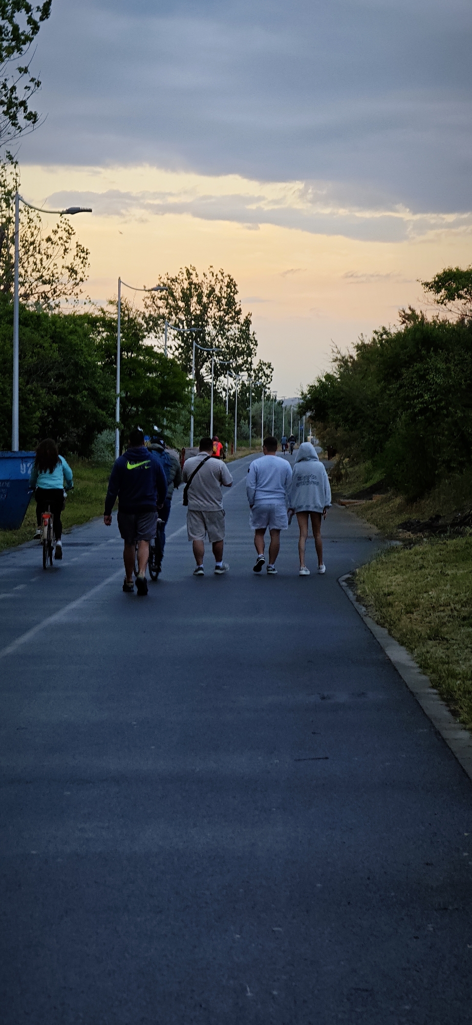 The image depicts a paved path in Burgas, Bulgaria, with a group of people moving away from the viewer towards the horizon. A person on a bicycle is on the left. The path is flanked by trees and streetlights. The sky shows a sunset.