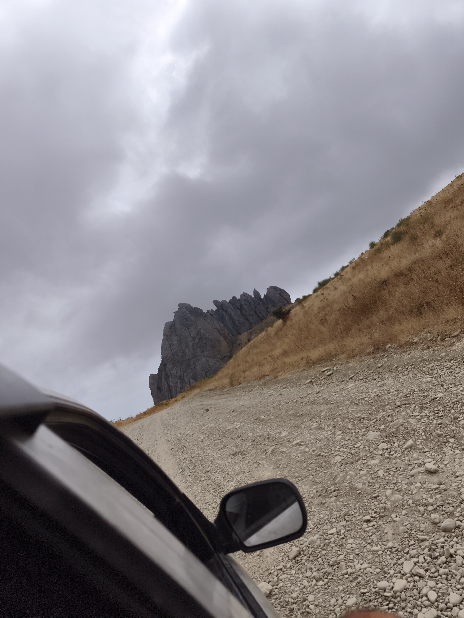 The image depicts a view from inside a car driving on a gravel road in Qalaşıxı, Azerbaijan. The road is strewn with pebbles and leads uphill towards a rocky outcrop. To the right of the road, a dry, grassy hillside slopes upwards, dotted with sparse green shrubs. In the distance, a jagged rock formation stands prominently against a cloudy, overcast sky. The side of the car, including the side mirror, is visible in the foreground, suggesting the perspective of someone traveling through this mountainous terrain.