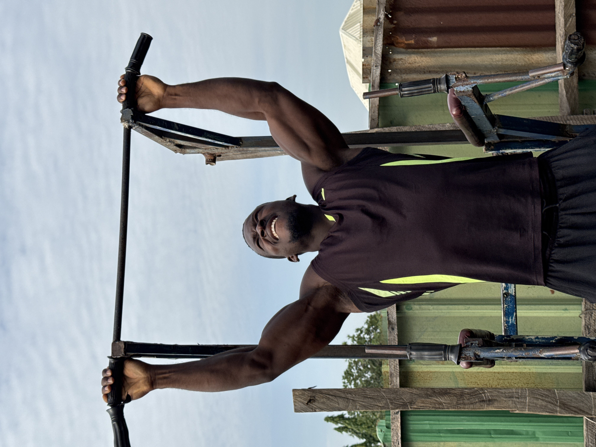 A Black male is performing a pull-up on a worn pull-up bar. He is wearing a dark sleeveless shirt with neon green accents and black shorts. His eyes are closed and he appears to be straining. The pull-up bar is part of a rusty, outdoor exercise apparatus. Behind him, there is a fence made of corrugated metal and some wooden structures. The sky is overcast. The image was taken in Akwanga, Nigeria.