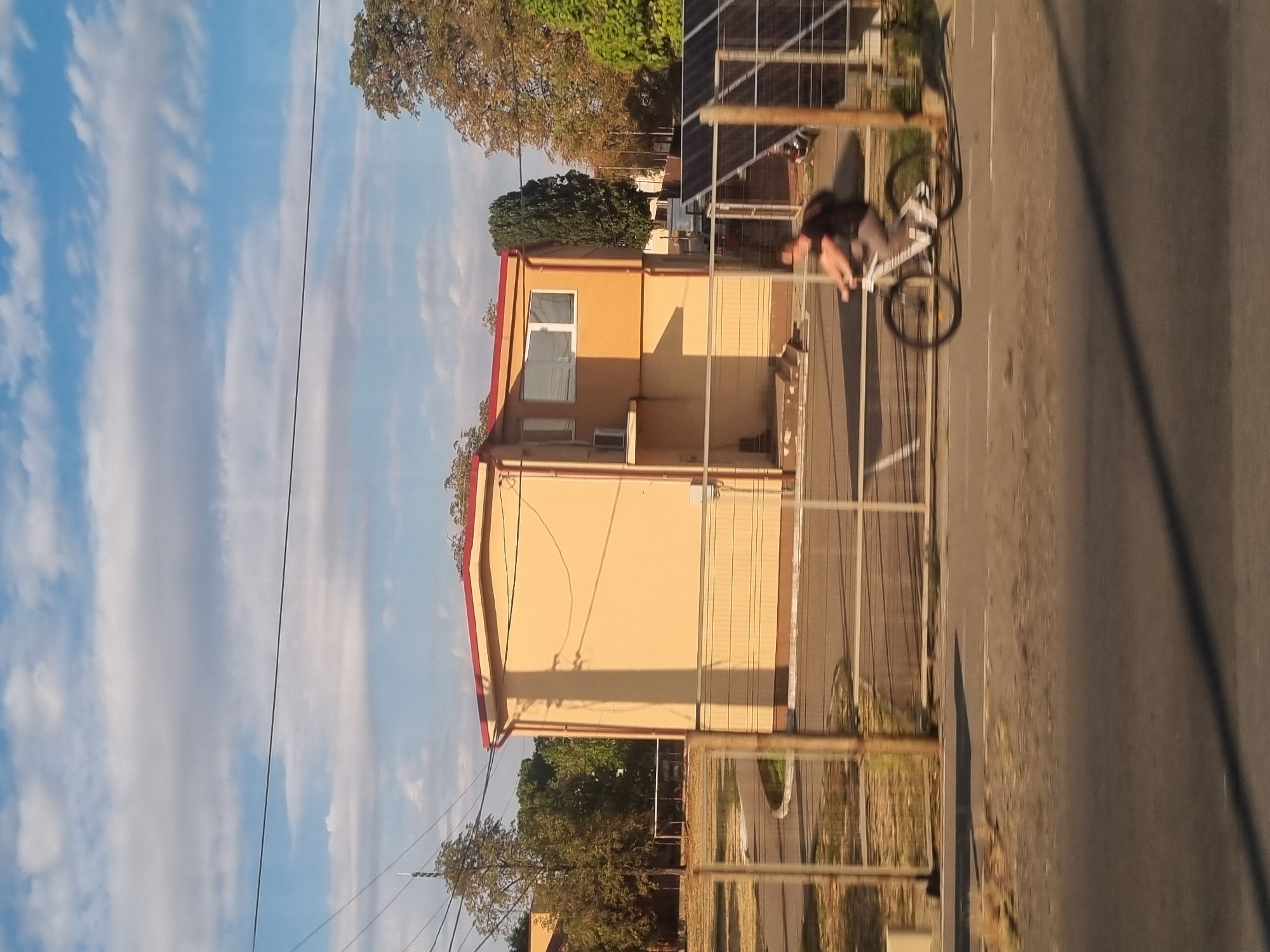 The image, taken in Sântion, Romania, depicts a suburban street scene under a partly cloudy sky. A person is riding a bicycle in the foreground, moving from left to right across the frame. The cyclist, wearing a dark t-shirt and grey pants, carries a backpack. Behind the cyclist is a metal fence, and beyond that, a light-yellow building with a red roof. The building has at least two windows visible on its facade, one of which is white-framed. To the right of the building, a tall, dark green tree stands out. In the far right of the image, a section of what appears to be solar panels is visible, also behind a fence. The ground in the foreground is paved, with a strip of dry grass and dirt to the left. The overall lighting suggests it is late afternoon or early evening, with shadows cast by the building and other objects.