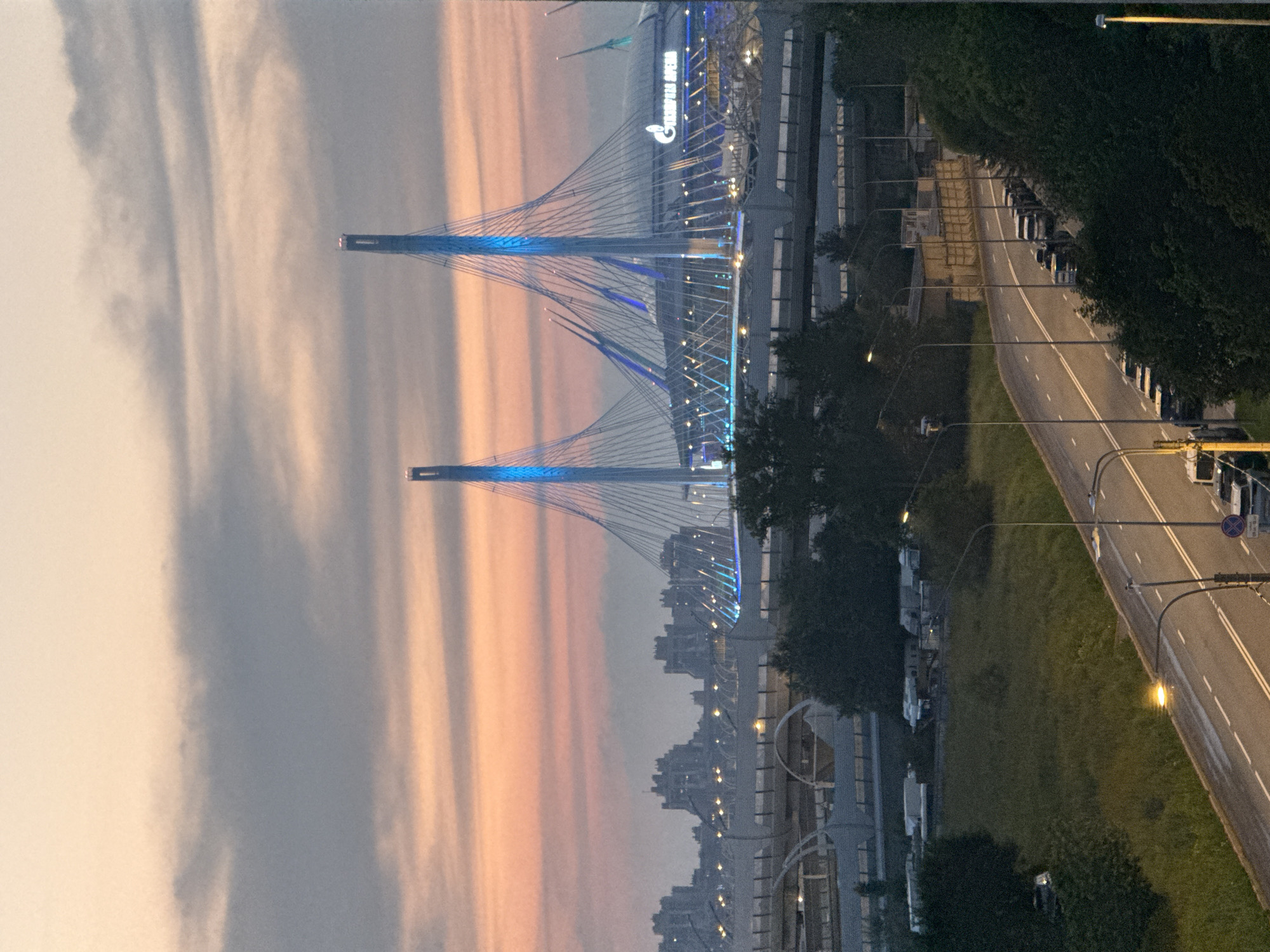 The image shows the Gazprom Arena in Saint Petersburg, Russia, at dusk. The stadium is illuminated with blue lights, and its distinctive cable-stayed roof is visible. In the foreground, there is a road with cars parked along the side and a grassy embankment. Several large buildings and structures, including bridges, are visible in the middle ground. The sky is a soft gradient of orange and gray, indicating the setting sun.