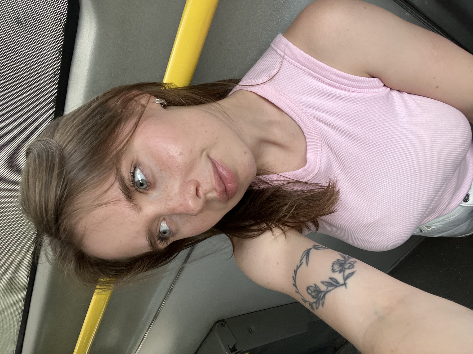 A young woman with light brown hair and blue eyes is sitting on a bus in Chaikovsky, Russia. She is wearing a pink ribbed tank top and white shorts. She has a floral tattoo on her left arm. The bus has a yellow pole and a grey seat. The woman is looking out the window.