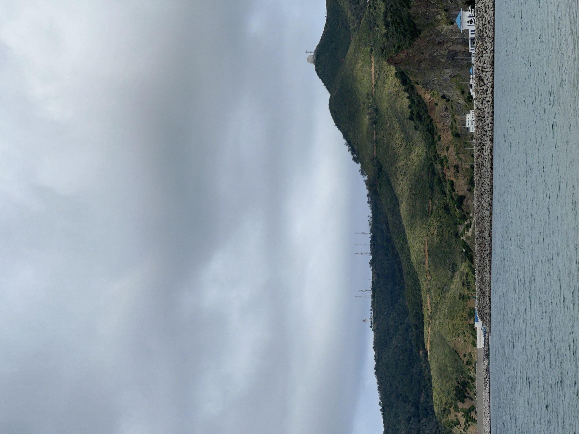 The image depicts a landscape in M'Diq, Morocco, featuring a coastline along the right edge of the frame. The water extends vertically. Along the shore, there's a breakwater comprised of interlocking concrete structures. Buildings, primarily white with blue accents, are situated between the breakwater and a steep, green mountain. The mountain rises along the middle of the frame and appears to have communication towers positioned along its ridgeline. A spherical radome is visible near the mountain's peak. The sky is overcast with varying shades of gray clouds occupying the majority of the frame on the left. The photo appears to be taken from a slightly elevated position looking towards the mountain and the sea.