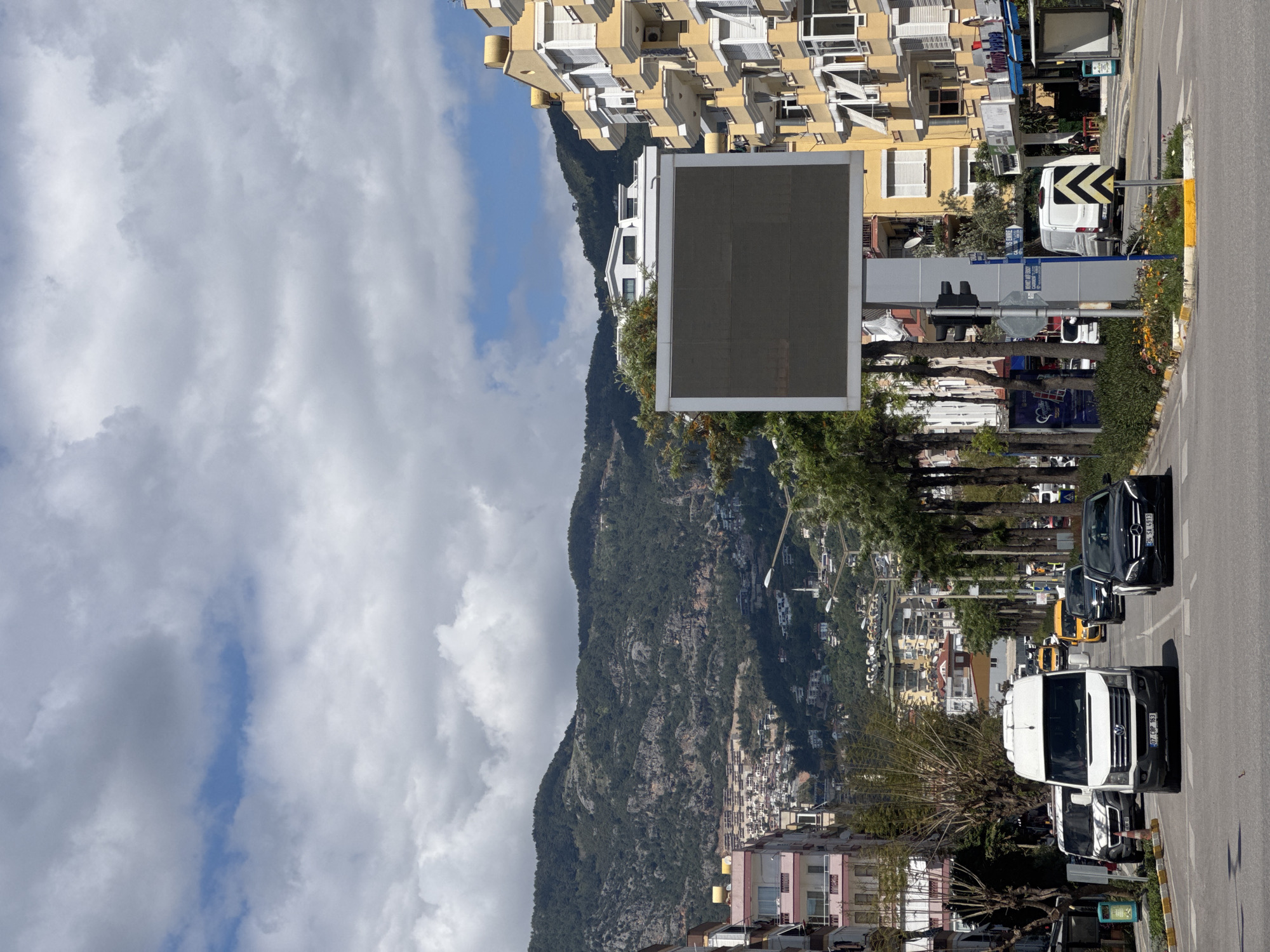A street scene in an unknown city, taken with the camera rotated 90 degrees counterclockwise, features a road with cars and buildings on either side, with a mountain backdrop partially obscured by clouds.
The road, oriented from bottom to top of the frame, has several vehicles parked along its edge including a large white van, a dark-colored sedan, and an orange construction vehicle.
On the left side of the road, at the bottom of the frame, is a building with pink and white facade. Above it is a mountain covered with vegetation. At the top of the mountain are buildings and structures.
On the right side of the road, near the top of the frame, are yellow buildings with balconies. Various signs and light poles are visible alongside the road.
The sky is predominantly covered with white clouds, with patches of blue visible through them.