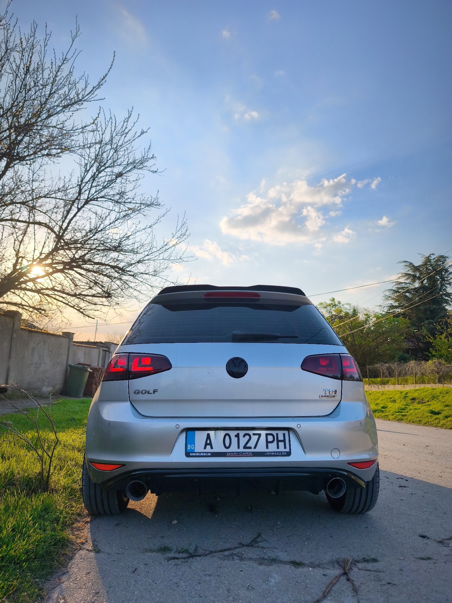 The image shows the rear of a silver Volkswagen Golf parked on a paved road in Avren, Bulgaria. The license plate reads "A 0127 PH" with the Bulgarian "BG" country code. The car features a black VW emblem and "GOLF" lettering on the left side, along with a "TDI Bluemotion" badge on the right. The brake lights are illuminated. The background includes a tree with bare branches on the left, through which the sun is partially visible, a concrete wall and a green trash bin. On the right, there is greenery and a wire fence. The sky is blue with scattered clouds.