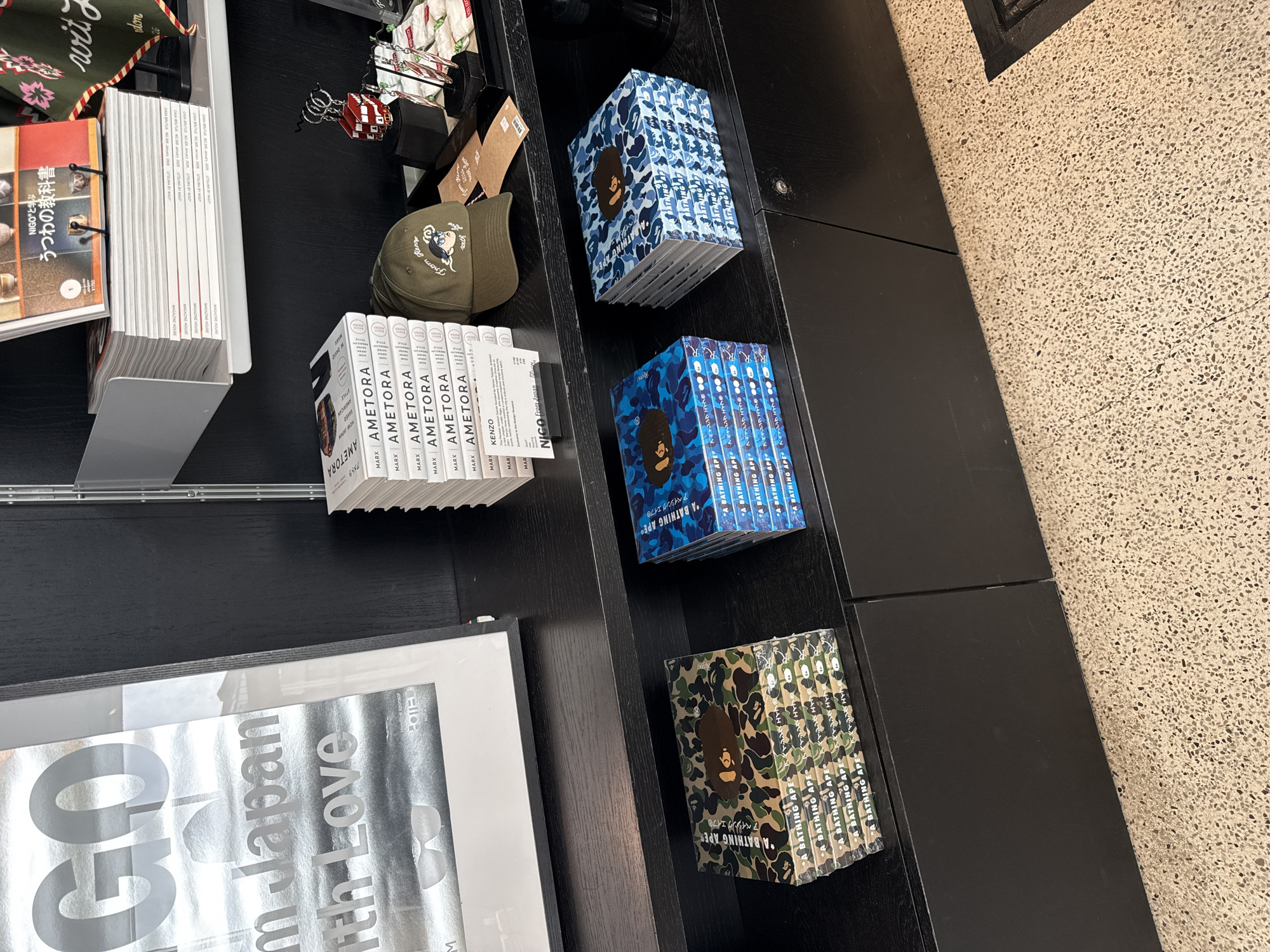 The image captures an indoor display, likely within a bookstore or gift shop in London, United Kingdom. The scene features several stacks of books and magazines arranged on dark, perhaps black, shelves or tables. The flooring is a light-colored terrazzo with small, dark speckled aggregate.
Prominently displayed are multiple copies of books with camouflage patterns. There are stacks of books in blue camouflage and others in green camouflage, all featuring the brand "A BATHING APE®" and its Japanese equivalent "アベイシングエイプ" printed on the spines and covers, often accompanied by the brand's iconic ape head logo.
Other stacks of books have white spines. One set is titled "AMETORA," with "MARX," "KENZO," "NOW JAPAN," and "NIGO From Japan" also visible on the spines. Another stack consists of magazines from "MAGAZINE HOUSE," specifically "CASA BRUTUS NO.308 January 2006 UTSUWA BY NIGO," and other editions. An open book with an orange/red cover shows the title "うつわの教科書" and "NIGOと学ぶ." In the foreground, a large, framed print or book cover features the text "GO in Japan With Love" with "POIE" visible near the bottom right.
Other objects include a dark green baseball cap embroidered with text that appears to read "from the world" or similar, placed next to the "AMETORA" books. Small decorative items, such as a red miniature house and a metallic wire sculpture, are also visible. A fabric item with a floral pattern hangs in the background.
No human activity is depicted, and the time of day is not discernible due to indoor lighting. The overall impression is a curated display of Japanese-themed literature and merchandise.
