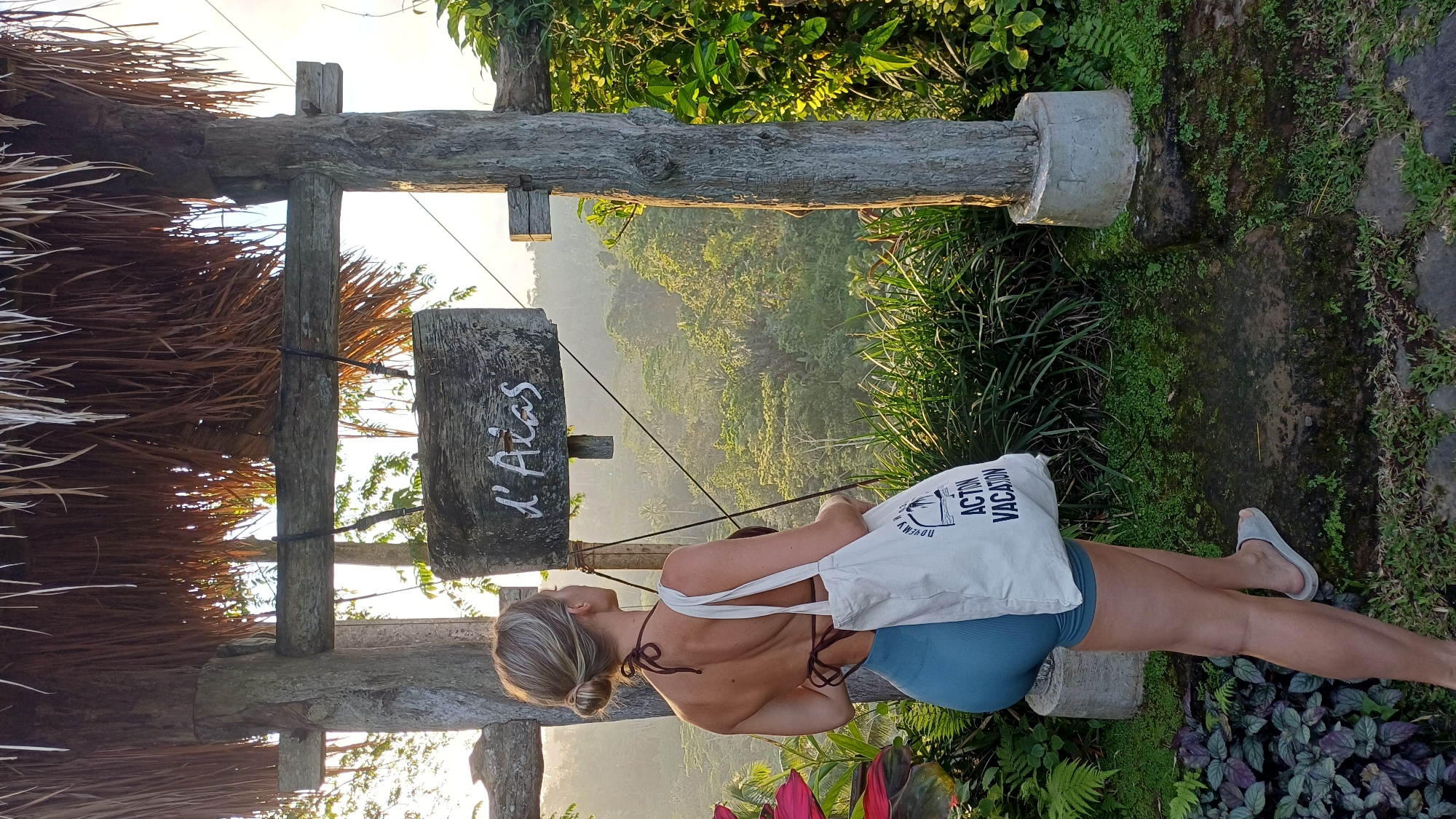 A back view of a person with fair skin, wearing a swimsuit and a white canvas bag, stands on a stone path with lush greenery and flowers around them. The path leads to a wooden structure with a thatched roof and a sign that reads "L'Alas." The scene is set in a valley with dense vegetation and a view of the horizon, possibly in the city of Tegallalang, Indonesia.