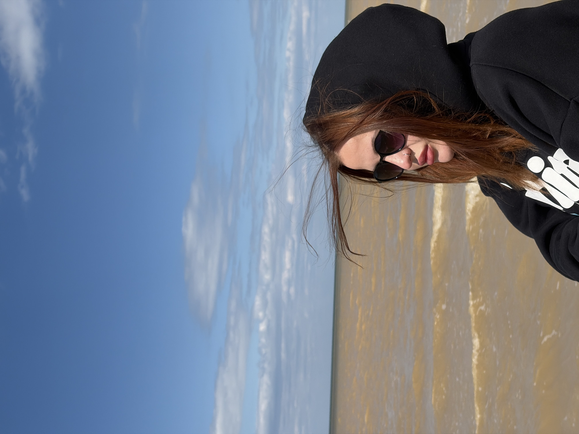 A person wearing a black hoodie and sunglasses is standing on a beach with light-colored sand or shallow water. The sky is blue with some clouds. The person's face is partially visible, and they appear to have brown hair. The image is possibly taken in Port-Katon, Russia.