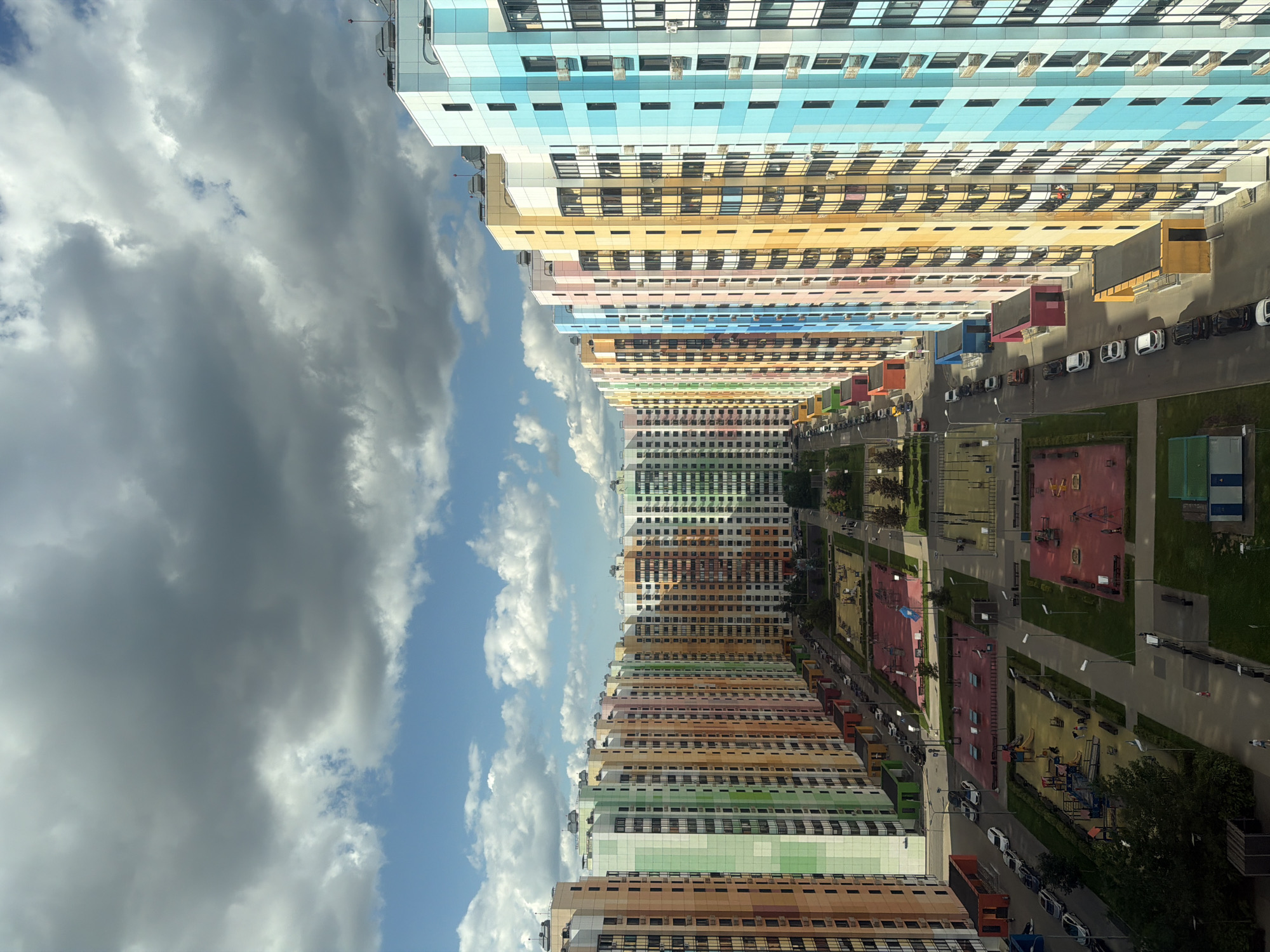 This is a high-angle aerial photograph of a residential complex in Moscow, Russia. The buildings are tall, multi-story apartment blocks painted in a variety of bright colors, including pastel green, orange, yellow, pink, and blue. The buildings are arranged in rows, forming a long, straight street lined with parked cars. In the foreground, there are several playgrounds with red and yellow surfaces, some with swings and slides. There are a few people visible on the playgrounds and sidewalks. The sky is partly cloudy with dramatic grey and white clouds against a bright blue sky. The overall impression is of a modern, colorful, and densely populated urban environment.
