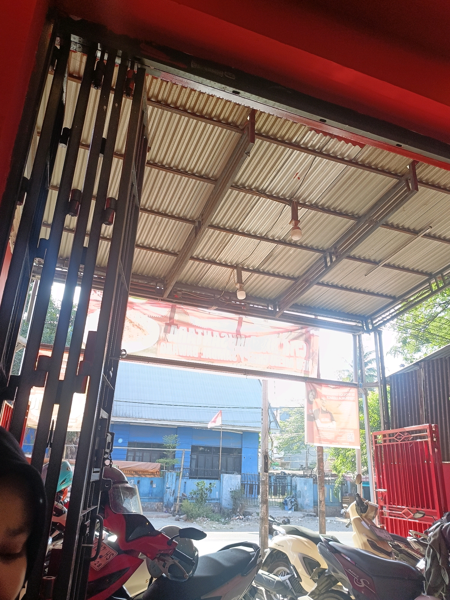 The image depicts a view from under a corrugated metal roof structure, likely an awning or covered area, looking outwards. The roof is supported by metal beams and has several light fixtures hanging from it. The scene is set in a street environment in Makassar, Indonesia.
In the foreground, to the left, a portion of a person's face and shoulder is visible, out of focus. In front of this person, and filling the lower part of the frame, are several parked motorcycles. One red motorcycle is prominent, with a black seat and silver accents. Other motorcycles, including a cream-colored one and a dark-colored one, are also visible.
Looking outwards, through a black metal gate or grill in the foreground, a street scene unfolds. A bright blue building is visible in the mid-ground, with multiple windows and a flagpole flying a red and white Indonesian flag. In front of the blue building is a light brown, tarp-covered structure or vehicle. Signs, possibly advertisements, are hanging from the roof structure, one of which features a person's image and text.
The environment appears to be an urban setting. Trees and other foliage are visible in the background, suggesting a warm climate. To the right, a red metal gate or fence is present. The overall impression is that of a street-level view, possibly from a shop or a residential area, with a covered waiting or parking area for motorcycles. The lighting suggests it is daytime, with sunlight filtering through the opening.
