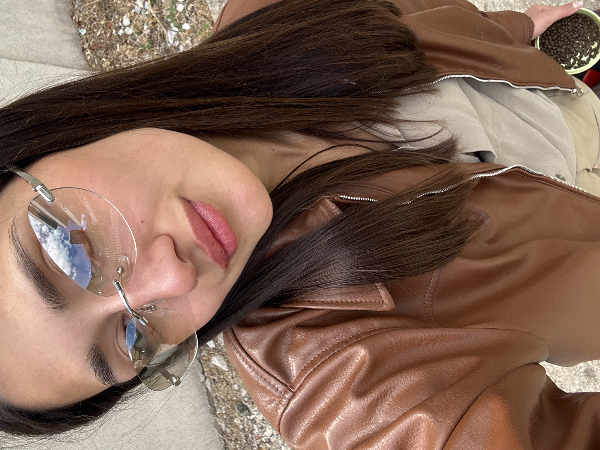 A woman with long brown hair wearing rimless glasses and a brown leather jacket is shown in a close-up, rotated portrait. She has pink lipstick and fair skin. A hand holding a green container filled with dark brown beans is visible on the upper right side of the frame. The background consists of gravel and plants. There are no identifiable indicators to confirm the location in Boreti, Montenegro.