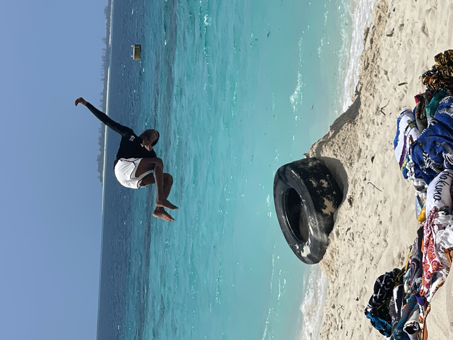The image displays an outdoor scene on a white sand beach in Zanzibar, Tanzania, under a clear blue sky. An adult male, with dark skin, is captured mid-air above the shallow, turquoise ocean water, performing an acrobatic flip. He is wearing a black long-sleeved rash guard and white shorts with a black stripe, and is barefoot. His body is horizontally oriented, with knees bent towards his chest and arms raised. On the white sand in the foreground, to the lower left, a pile of colorful, patterned textiles, including towels (one visibly marked "Hakuna"), is present. To the right of the textiles, a large, black, weathered vehicle tire is partially buried in a sand mound at the water's edge. The ocean water exhibits a clear transition from light turquoise near the shore to a deeper blue further out. A yellow rectangular buoy floats in the distant water. Low-lying landmasses or islands with sparse vegetation are visible on the distant horizon.