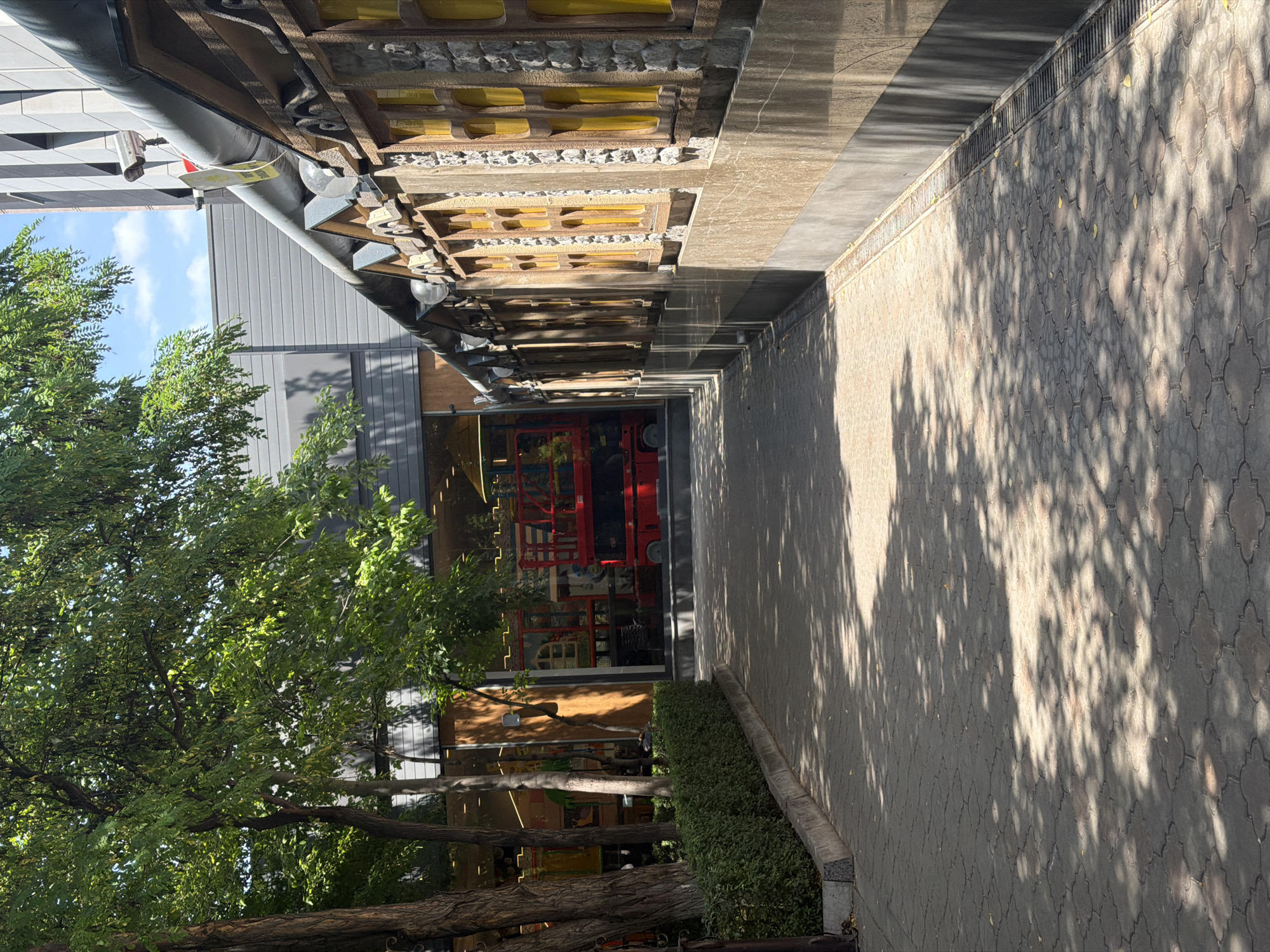 A view of a paved walkway in Yerevan, Armenia, adjacent to a building with decorative windows and an overhanging roof. Trees with green foliage cast dappled shadows on the walkway. The building has a textured facade with stone-like elements and windows that appear to be filled with yellow material. To the left, a low, manicured hedge lines the walkway, and beyond it, a building with large glass windows is visible, revealing interior structures and a red scissor lift. The sky above is blue with some clouds.