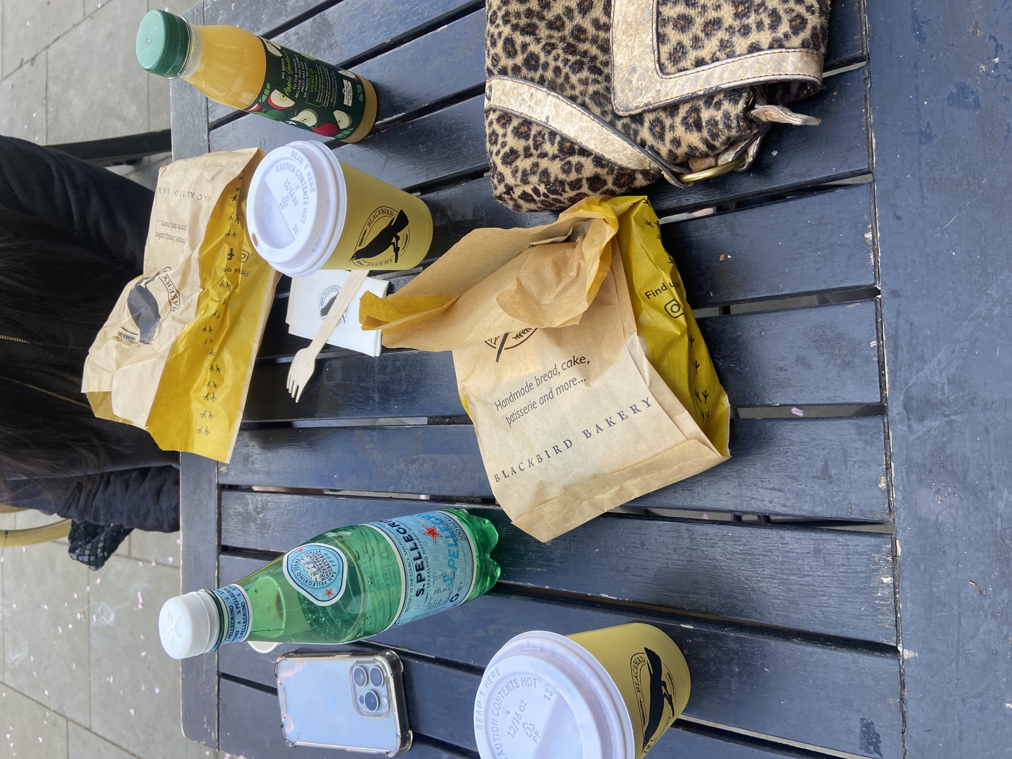 A cluttered black picnic table presents remnants of a meal purchased from Blackbird Bakery. A leopard print handbag is on the table along with a bottle of San Pellegrino, a juice bottle, two Blackbird Bakery coffee cups, a Blackbird Bakery pastry bag, a brown takeout bag, a cellphone, and disposable wooden cutlery. The table is in an outdoor setting, possibly in London, United Kingdom, given the presence of Blackbird Bakery, which has locations in London. A person is partially visible to the left. There are flower petals on the picnic table.
