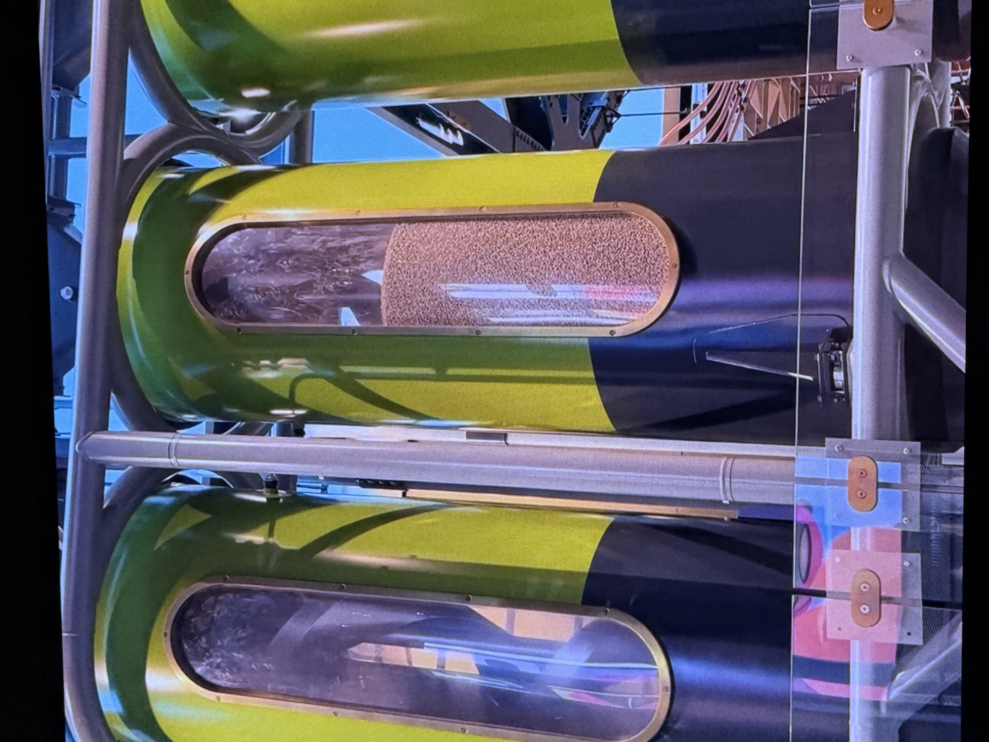 The image shows a close-up view of a large industrial machine, likely a grain silo or storage system, with three vertical cylindrical tanks visible. The tanks are painted in a bright lime green with a dark navy blue base and feature prominent oval-shaped windows made of glass or clear plastic. The window on the left tank is partially obscured by reflections, but appears to contain some solid material. The middle tank's window is filled with a dense collection of small, light-brown pellets or grains. The third tank to the right is mostly visible and appears empty. The tanks are supported by a metal framework, and pipes run horizontally above them. The background is blurred but suggests an outdoor environment with a bright blue sky and some industrial structures. The image was captured in Dubai, United Arab Emirates. There are no people visible in the frame.