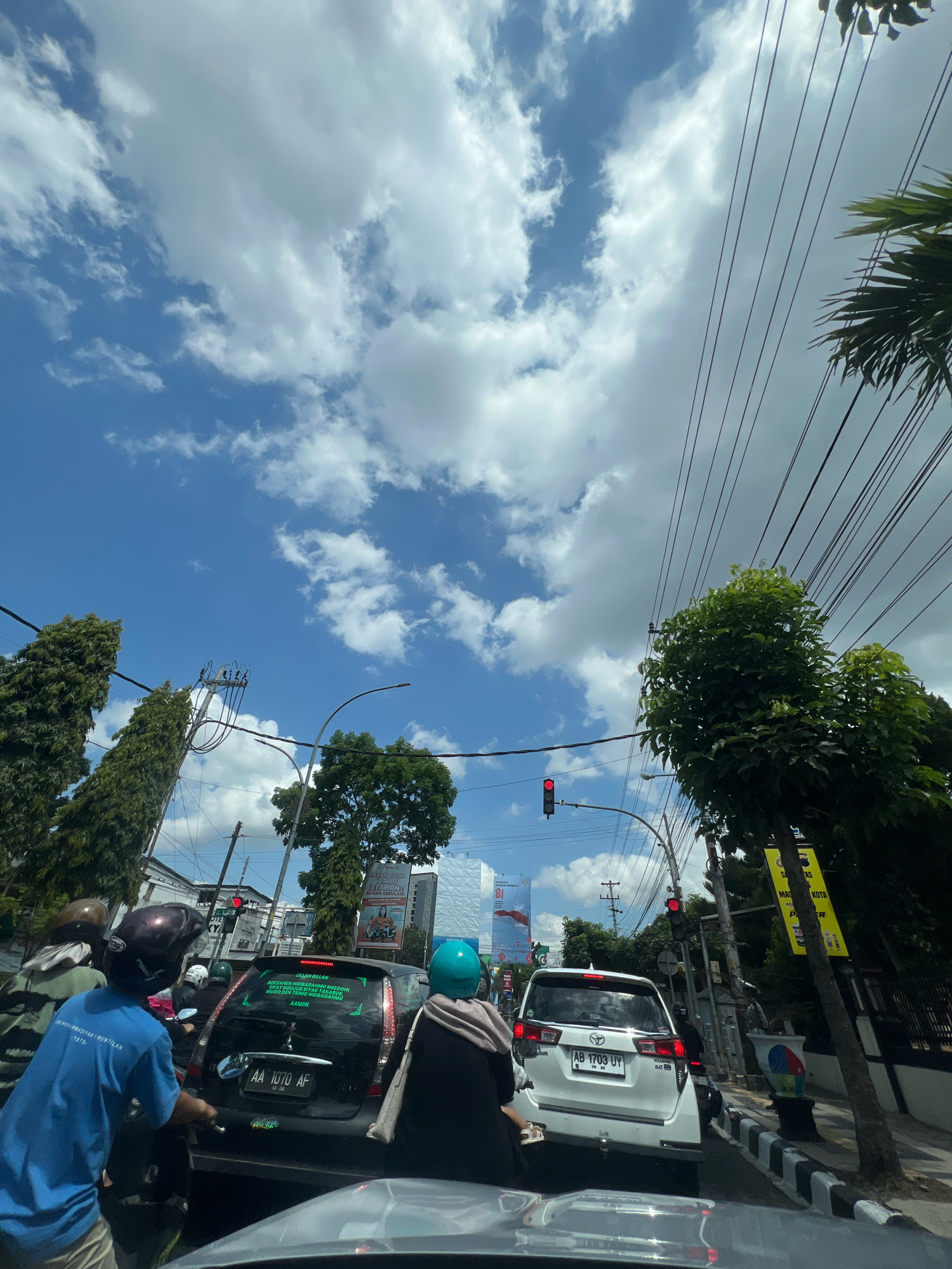 This is a street-level, medium-shot, eye-level photograph taken in the city of Magelang, Indonesia, during daylight hours. The scene depicts a traffic jam on a road, likely an intersection given the presence of traffic lights. The sky is blue with scattered white clouds, and several power lines are visible crisscrossing the frame overhead. Lush green trees line the sides of the road, with some tall, slender trees on the left and a thicker, leafier tree on the right.
In the foreground and midground, numerous vehicles are stopped. On the left, a person wearing a blue shirt and a helmet is on a motorcycle. To their right, a black car with a license plate ending in "AF" is stopped, and text in green is visible on its rear window. Further back, a white SUV with a license plate ending in "UY" is also stopped. Several other motorcycles and cars are present in the traffic jam, with people wearing helmets. The traffic lights are red, indicating that traffic is halted.
In the background, tall buildings are visible, suggesting an urban or semi-urban environment. A tall billboard with red and white graphics is also present on the left side of the road. The road surface is paved, and a concrete barrier with black and white stripes runs along the right side of the road, separating it from a sidewalk or embankment. The overall impression is a typical busy day in an Indonesian city, with a mix of modern infrastructure and abundant greenery.