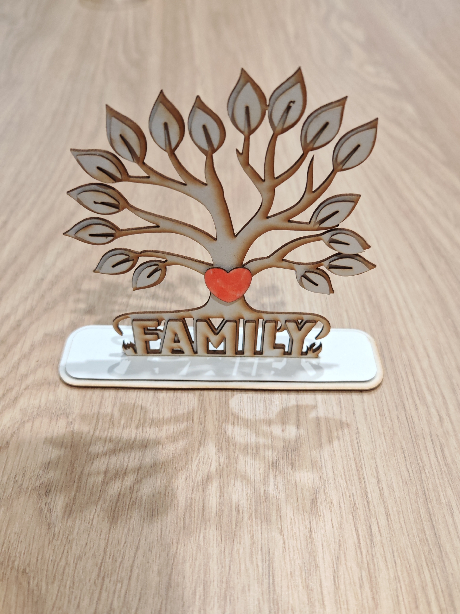 The image shows a close-up, eye-level shot of a wooden family tree ornament. The ornament is laser-cut and features a central tree with multiple branches and leaves. A small, red heart is positioned on the trunk of the tree. The word "FAMILY" is cut out of wood and forms the base of the ornament, resting on a white rectangular platform. The entire piece sits on a light brown, wooden surface that appears to be a table or desk. The lighting creates a shadow of the ornament on the wooden surface, indicating an overhead light source. The image is taken indoors, with no visible elements indicating a specific location in Plovdiv, Bulgaria.