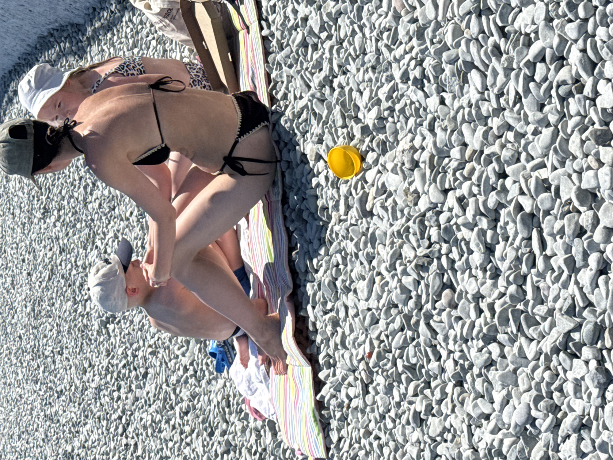 This media file shows a group of people on a pebble beach, likely in Partenit, Ukraine. The image is taken from a high-angle perspective looking down at the subjects. The scene is bathed in sunlight, suggesting it is daytime with clear weather.
There are at least three people visible. One person, likely an adult, is wearing a black bikini and a green baseball cap. They are bending down, facing towards another person. This second person, possibly a child, is wearing a white sun hat and a leopard-print bikini top. They are also bending down, looking towards the first person. A third person, also appearing to be a child, is wearing a white cap and is partially visible, looking down. A striped beach towel is spread out on the pebbles beneath the people. A small yellow object, possibly a toy or lid, is visible on the pebbles to the right of the people. The ground is covered in smooth, light-colored pebbles.
The people appear to be engaged in a casual activity on the beach, possibly playing or interacting with each other. The overall mood seems relaxed and summery. There are no visible text elements in the image.