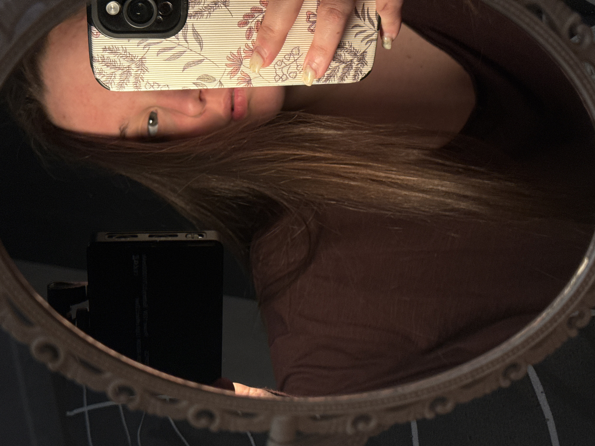 A woman is taking a selfie in a round, ornate mirror. Her face is partially obscured by her phone, which has a floral patterned case. Her hair is long and brown. She is wearing a brown shirt. There is a black rectangular object partially visible behind her phone. The background appears to be a dark, possibly striped surface. There is no information to confirm the location.