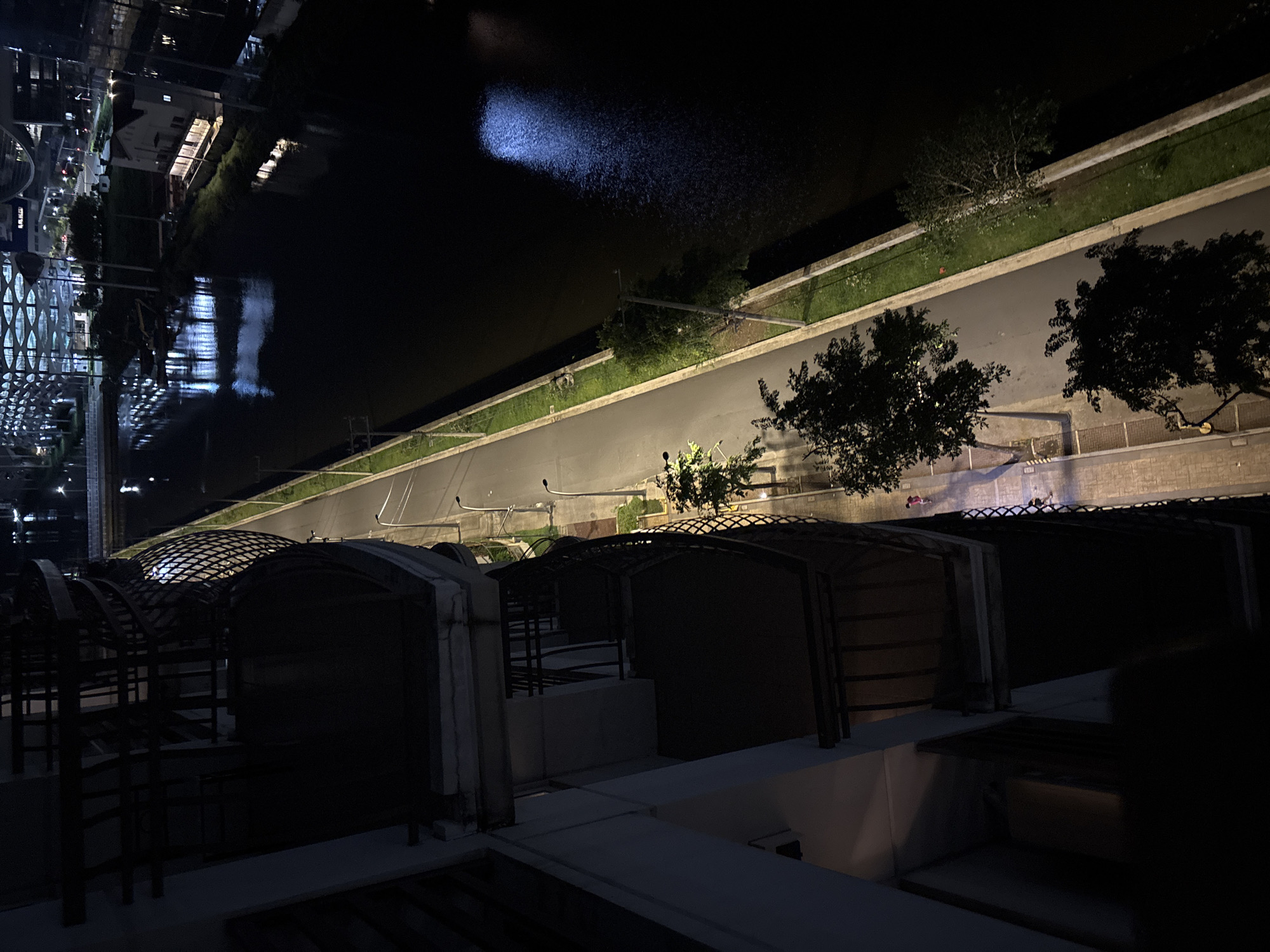 The image shows a night view from a balcony in Pasay City, Philippines, looking down at a canal, a pedestrian walkway, and a road. The perspective is from a high-rise building, with the dark facade and railing of the balcony dominating the left side of the frame. The canal, on the right, is dark and reflects the lights of surrounding buildings and streetlights, creating a shimmering, almost ethereal effect on its surface.
A narrow, paved walkway runs along the edge of the canal, lined with a strip of green grass and intermittent trees. Streetlights are visible along this walkway, casting pools of light. The road is parallel to the walkway, also illuminated by streetlights.
Further in the distance, across the canal, are modern buildings with illuminated windows and architectural details. One building, in particular, has a distinctive grid-like facade. There are also some indistinct structures and lights suggesting the presence of other urban infrastructure.
In the foreground, on the walkway, a single person is visible, wearing a pink top, walking away from the camera. Another figure, possibly a child, is seen further down the walkway. The overall scene depicts a quiet urban night, with the interplay of darkness and light creating a dramatic atmosphere.