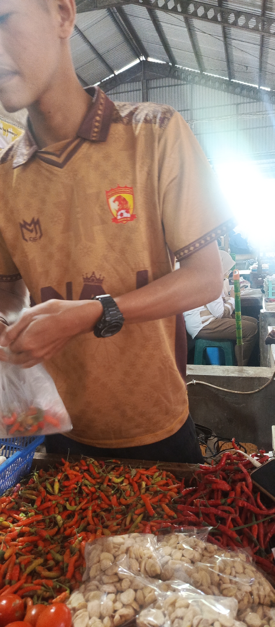 A person wearing a tan-colored jersey with a maroon collar and sleeve trim stands in what appears to be an outdoor market in Malang, Indonesia. The jersey has a crest on the left breast and a logo on the left sleeve. The person is holding a clear plastic bag filled with small red chilies. In front of the person are piles of red chilies and some packaged goods that appear to be nuts or seeds. Behind the person, other market stalls and people are visible, with a corrugated metal roof structure overhead suggesting an indoor or covered market. The lighting indicates it is daytime, possibly with bright sunlight filtering through.