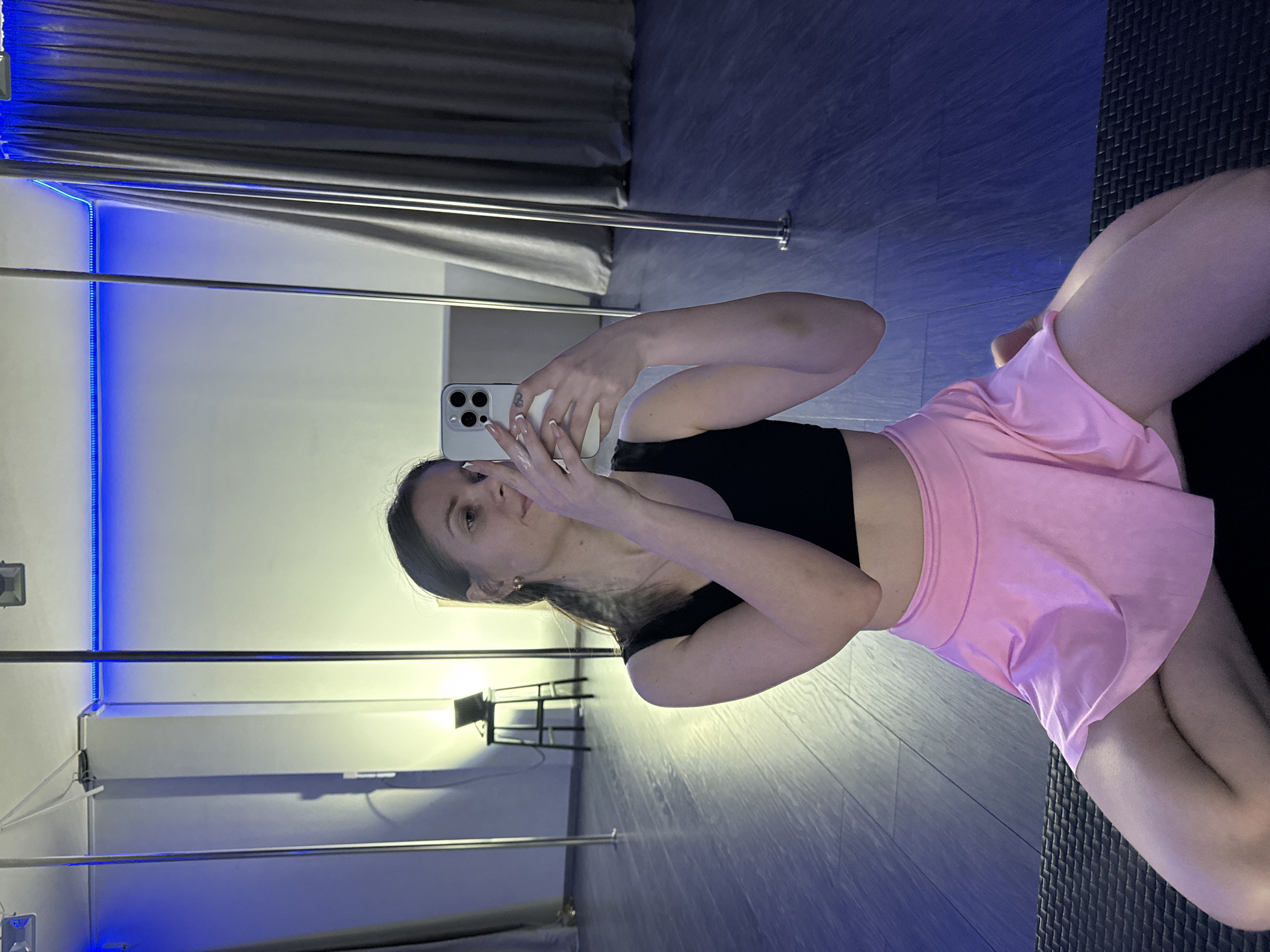 A woman is taking a selfie in what appears to be a dance or exercise studio. She is wearing a black crop top and a pink skirt, and she is holding a smartphone. The studio has hardwood floors, a mirror, and dance poles. The lighting is a mix of natural and artificial, with blue LED strips lining the ceiling. A metal chair is visible in the background. The location appears to be indoors, possibly in Batumi, Georgia. The presence of dance poles suggests the studio is used for pole dancing or a similar activity.