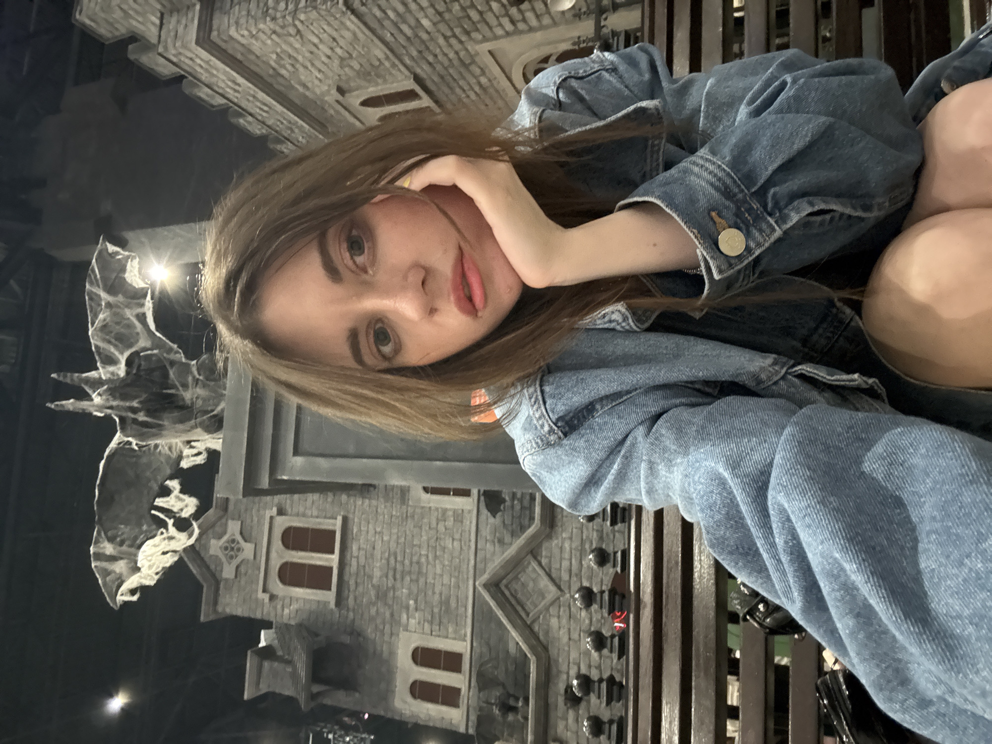 A young woman with light brown hair is posing for a selfie outdoors, resting her head on her hand and looking at the camera. She is wearing a denim jacket. Behind her, a decorative facade of a gothic-style building is visible. The scene is located in Moscow, Russia.