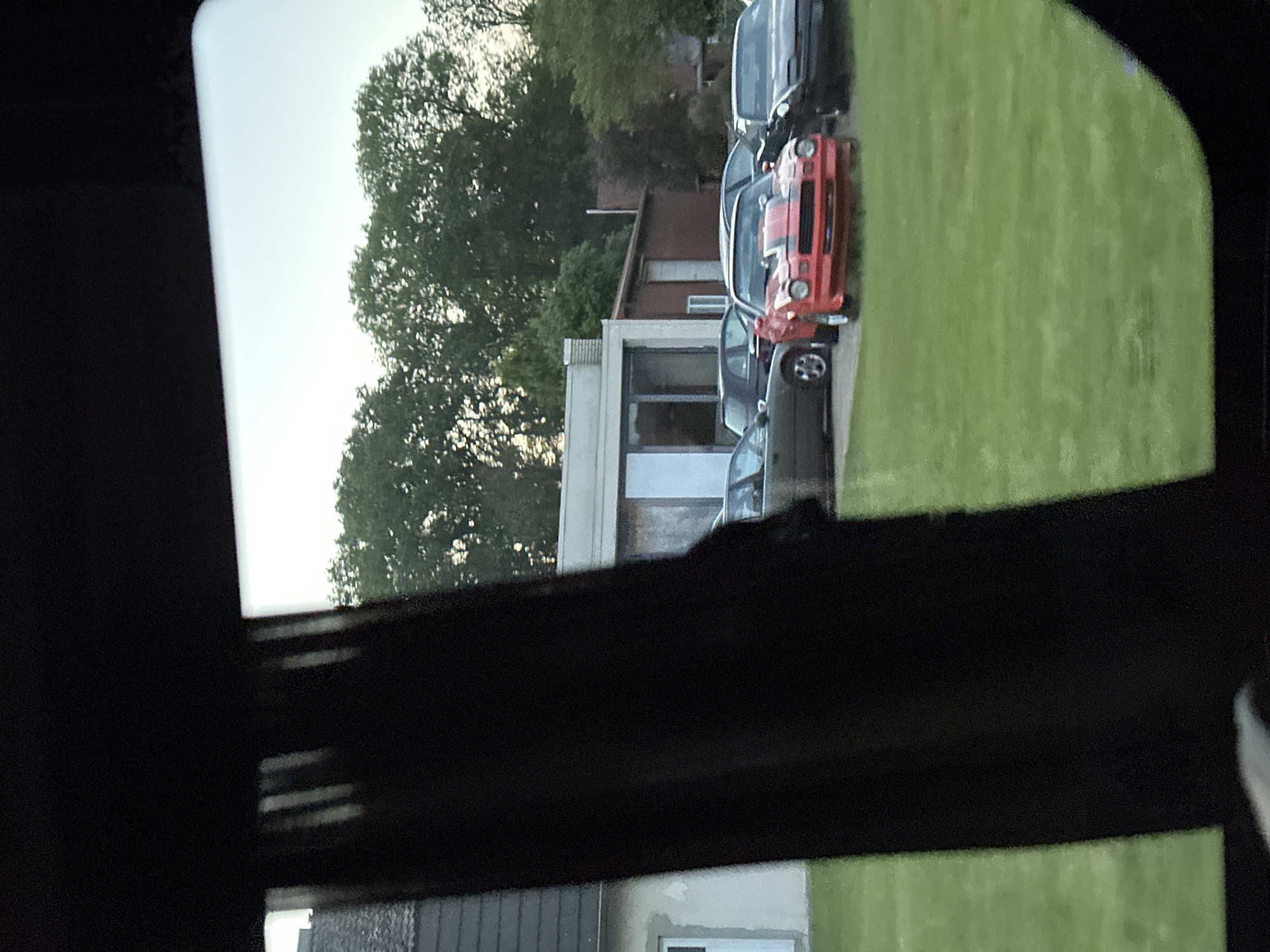 The image shows an outdoor scene, possibly in Harsefeld, Germany, viewed through a dark window frame. A light-colored building with a chimney and a darker brick building behind it are visible. Several cars are parked on a grassy area. One appears to be an older model orange/red sports car with black stripes. The day is overcast. Trees with dense foliage are present in the background. A building with a gray roof and a small window is visible in the lower foreground, outside the window frame.