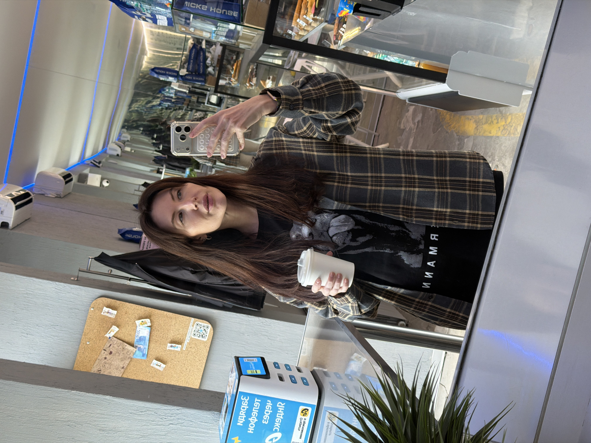 A woman takes a selfie in front of a mirrored display at a location in Perm, Russia, as indicated by the text on a nearby payment kiosk. She is holding a white cup with a lid in her left hand and using a smartphone to take the picture with her right hand. She is wearing a plaid blazer over a black t-shirt with white lettering. Her long brown hair falls over her shoulders. To her left is a corkboard with pinned notes and a coat hanging below. The background features a food display case and mirrored surfaces, reflecting the interior of the establishment. A potted plant is visible near the payment kiosk.