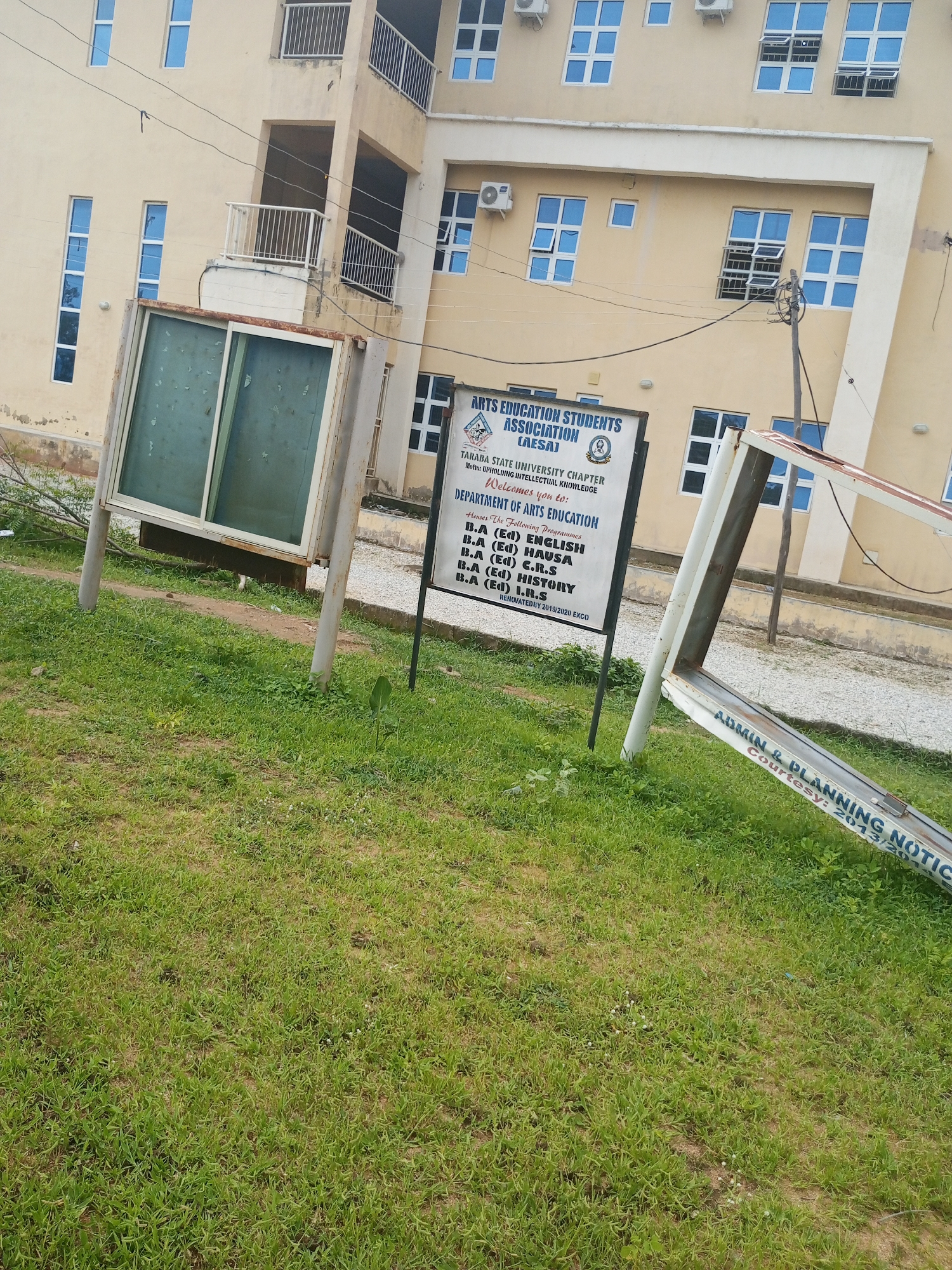 This media file captures an outdoor scene in Jalingo, Nigeria, likely on a bright, possibly sunny day given the lighting. The focus is on a section of a pale yellow building with multiple floors and many windows, some open and some closed. The building appears to be a university or institutional structure.
In the foreground, a grassy area with patches of dirt is visible. A leaning, metal-framed signpost lies on the grass, with some text still legible on it, including "ADMIN & PLANNING NOTICE" and "Courtesy: 2013/201". To the left of this, a larger, upright signpost stands, also with visible text. This sign advertises the "ARTS EDUCATION STUDENTS ASSOCIATION (AESA)" of "TARABA STATE UNIVERSITY CHAPTER" and lists the programs offered by the "DEPARTMENT OF ARTS EDUCATION," such as "B.A (Ed) ENGLISH" and "B.A (Ed) HISTORY." The motto of the association, "UPHOLDING INTELLECTUAL KNOWLEDGE," is also present.
Another large, framed display case with a glass front is positioned on the left side of the image, near the grass. The glass is somewhat obscured, possibly due to age or weather.
The overall scene suggests a university campus environment. The presence of the signs and the building architecture indicate an academic setting. The leaning signpost implies some recent disturbance or perhaps just the passage of time affecting the structure. The weather appears to be clear, allowing for the ample daylight.