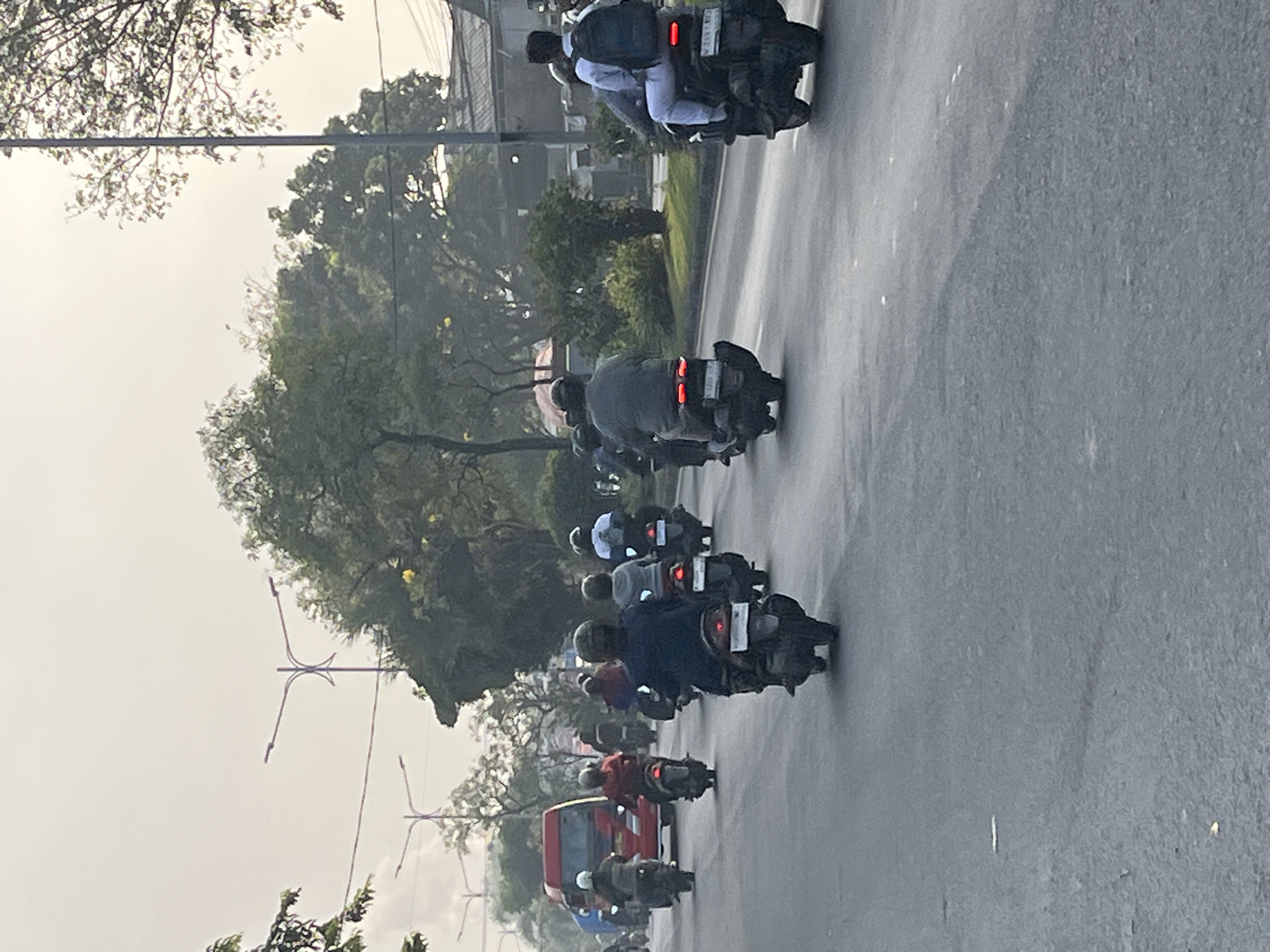 The image depicts a street scene in Gelam, Indonesia, with multiple motorcyclists traveling on a paved road under an overcast sky. The focus is on the flow of traffic, with several individuals riding motorcycles in the same direction. The environment suggests an urban or suburban setting with trees lining the sides of the road and streetlights visible. The vehicles are varied in color and design, and the people are wearing casual clothing and helmets. The overall impression is a snapshot of everyday life in Indonesia, capturing the common mode of transportation and the general atmosphere of the location.
