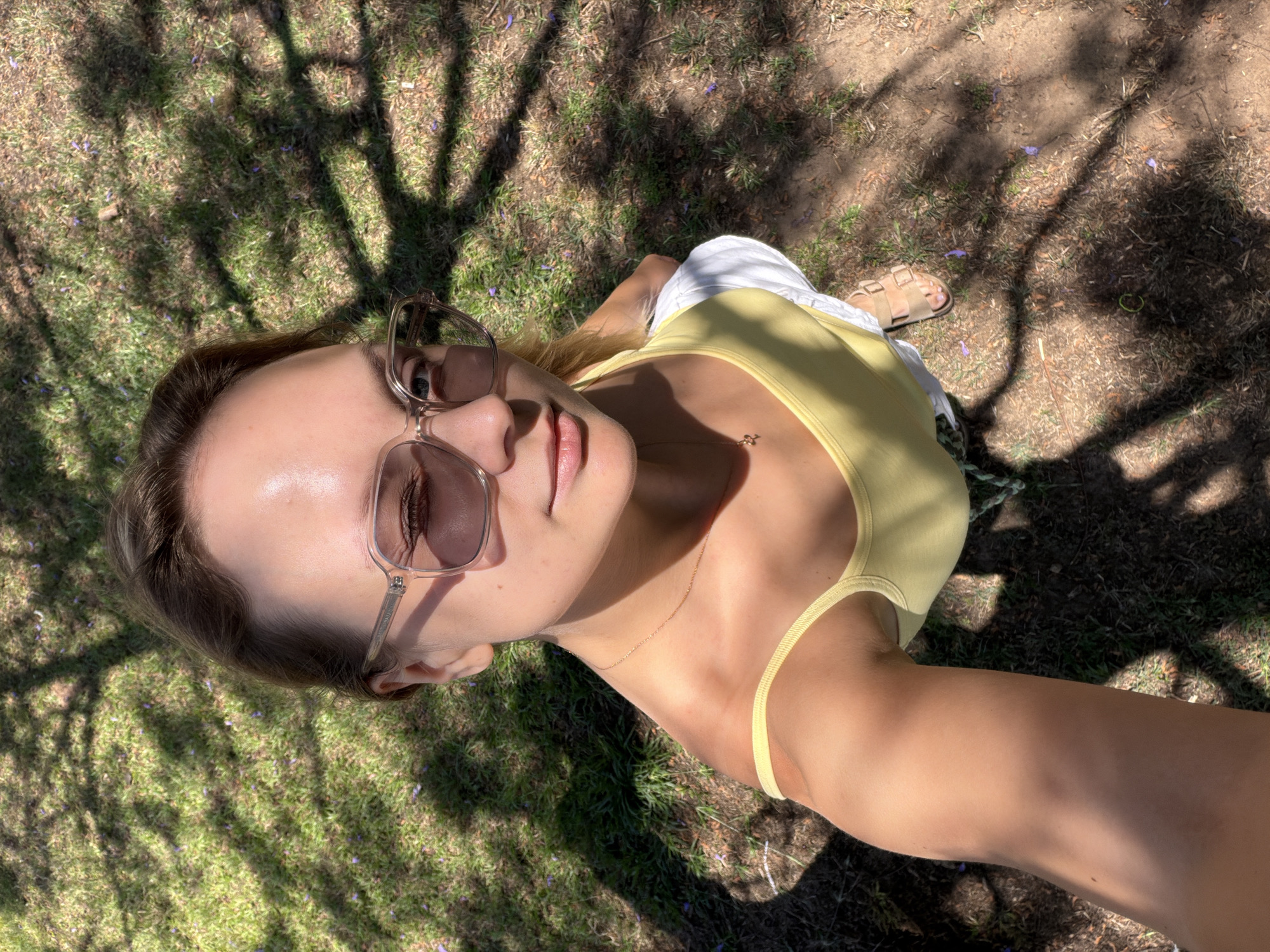 A high-angle, full-body selfie is taken by a young woman standing outdoors on a sunny day. She is wearing a yellow tank top, white shorts, and tan sandals. She has on oversized sunglasses and her hair is pulled back. The woman is looking up and to the side, with her left eye closed in a wink. She is holding the camera with her left hand, which is partially visible in the bottom left of the frame. The background consists of grass, dirt, and tree shadows, suggesting a park or garden setting. The presence of small purple flowers scattered on the ground might indicate a jacaranda tree. The scene appears to be located in Tel Aviv, Israel, given the context of the image.
