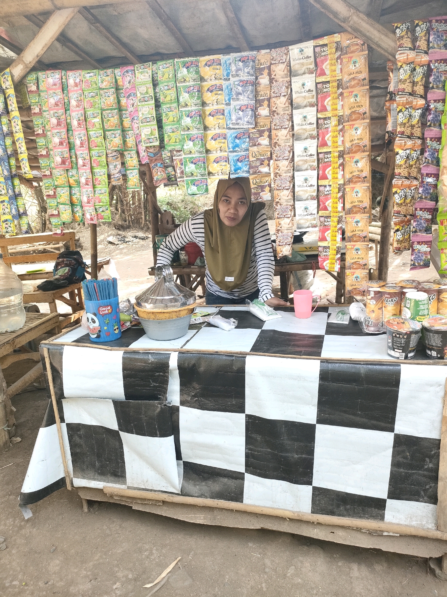 A woman wearing a brown headscarf and a striped shirt is standing behind a stall in Tongas Kulon, Indonesia. The stall has a checkered black and white tablecloth. Behind her, the stall is decorated with rows of packaged snacks and drinks. To the left of the woman, there is a large bottle of water and a blue cup filled with straws. To her right, there are several instant noodle cups and other packaged goods. The stall appears to be outdoors, with a wooden structure supporting a roof made of corrugated material. The ground around the stall is dirt.