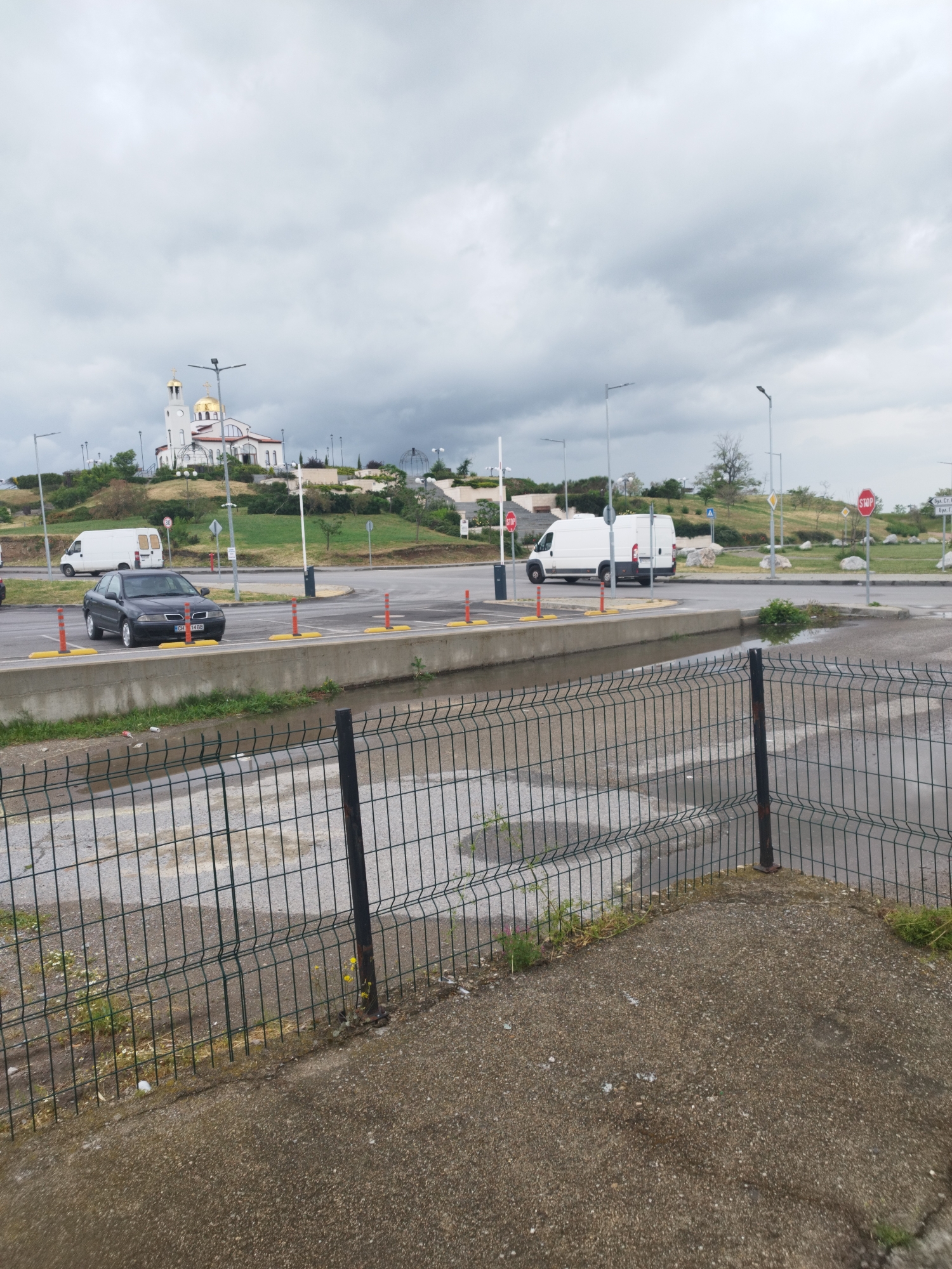 The image shows a parking lot in Unknown, Unknown, on an overcast day. A black car is parked in a designated spot. Several white vans are also parked in the lot. A modern church with gold domes is visible on a hill in the background. A drainage ditch runs between the parking spots and the foreground, with a chain-link fence separating the ditch from a concrete area in the front of the image. Streetlights are positioned throughout the parking lot and surrounding area. Stop signs are visible at intersections.