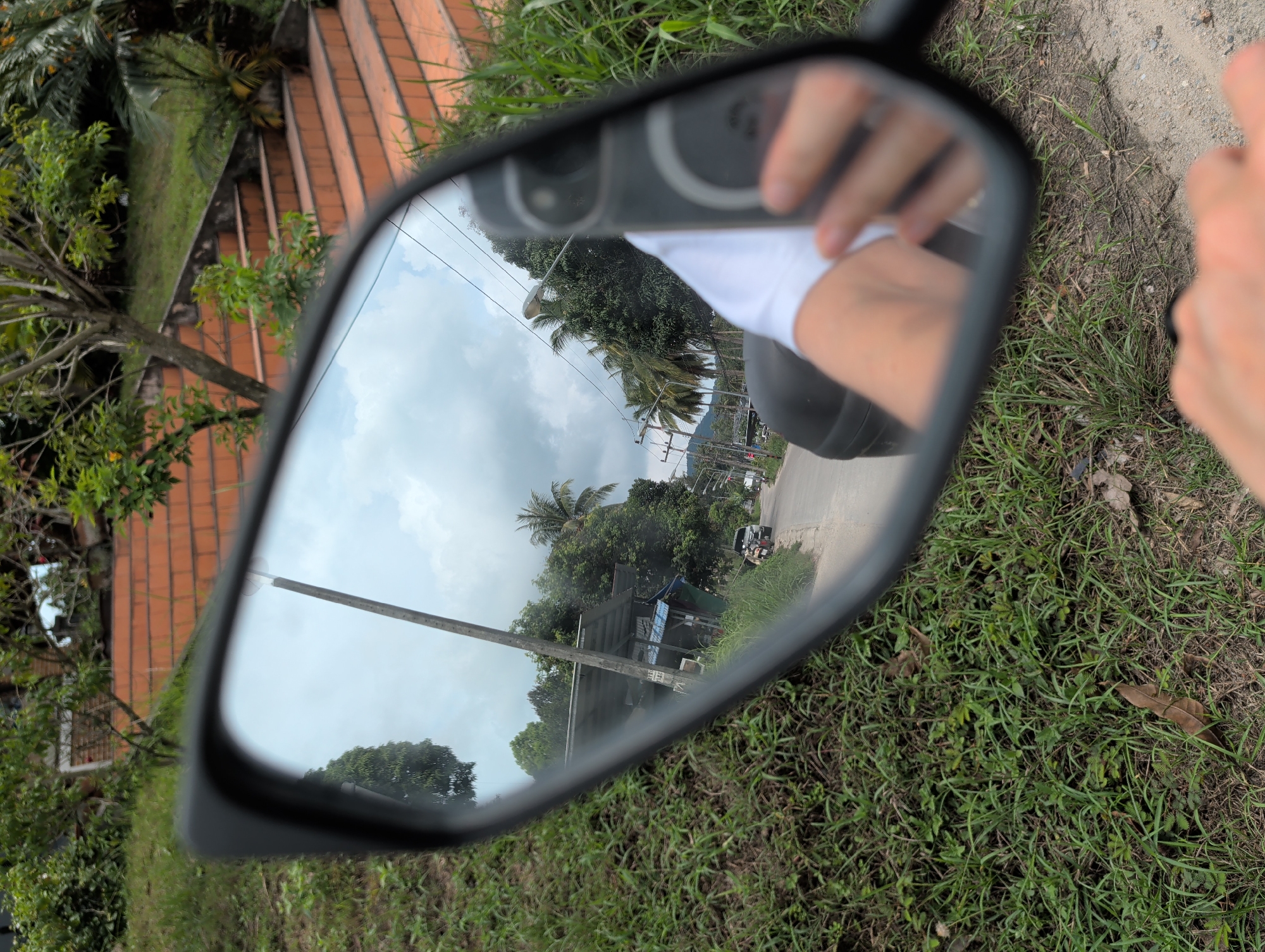 The image shows a motorcycle mirror reflecting a road in Moo 4, Thailand. The reflection includes a view of the road, trees, utility poles, buildings, and the sky. The person taking the picture and holding their phone is also visible in the mirror. The foreground surrounding the mirror consists of grass and dirt.