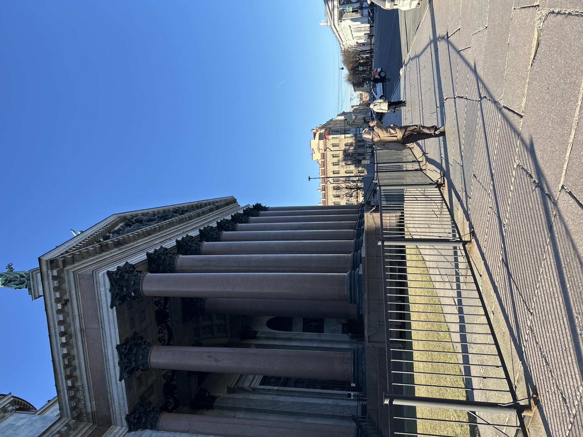 The image depicts a view of St. Isaac's Cathedral in St. Petersburg, Russia. The cathedral's imposing columns are visible on the left side of the frame, extending vertically towards the clear blue sky. A metal railing runs along the edge of a stone platform or sidewalk in the foreground. A person is standing near the railing, dressed in a tan jacket and dark pants, facing away from the camera. In the background, across the street, are other buildings, including a light-colored one with multiple windows. People and cars are visible on the street. The sunlight casts long shadows across the pavement.
