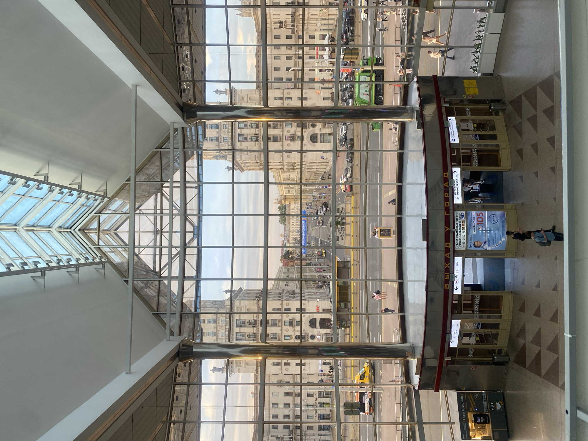 The image shows a view from inside a modern building with a large glass facade overlooking a street in Minsk, Belarus. The interior features a high, angled glass ceiling and large, polished metal pillars. In the foreground, there are two entrances to a metro station, indicated by signage in Cyrillic. People are visible entering and exiting the metro and walking outside on the street. The street scene includes several buildings with classical architecture, a yellow bus, a green garbage truck, and various cars. The overall impression is of a busy urban environment with a blend of modern and historical architecture. The specific location is the exit of the "Ploshcha Peramogi" (Victory Square) metro station in Minsk, Belarus, as indicated by the signage.