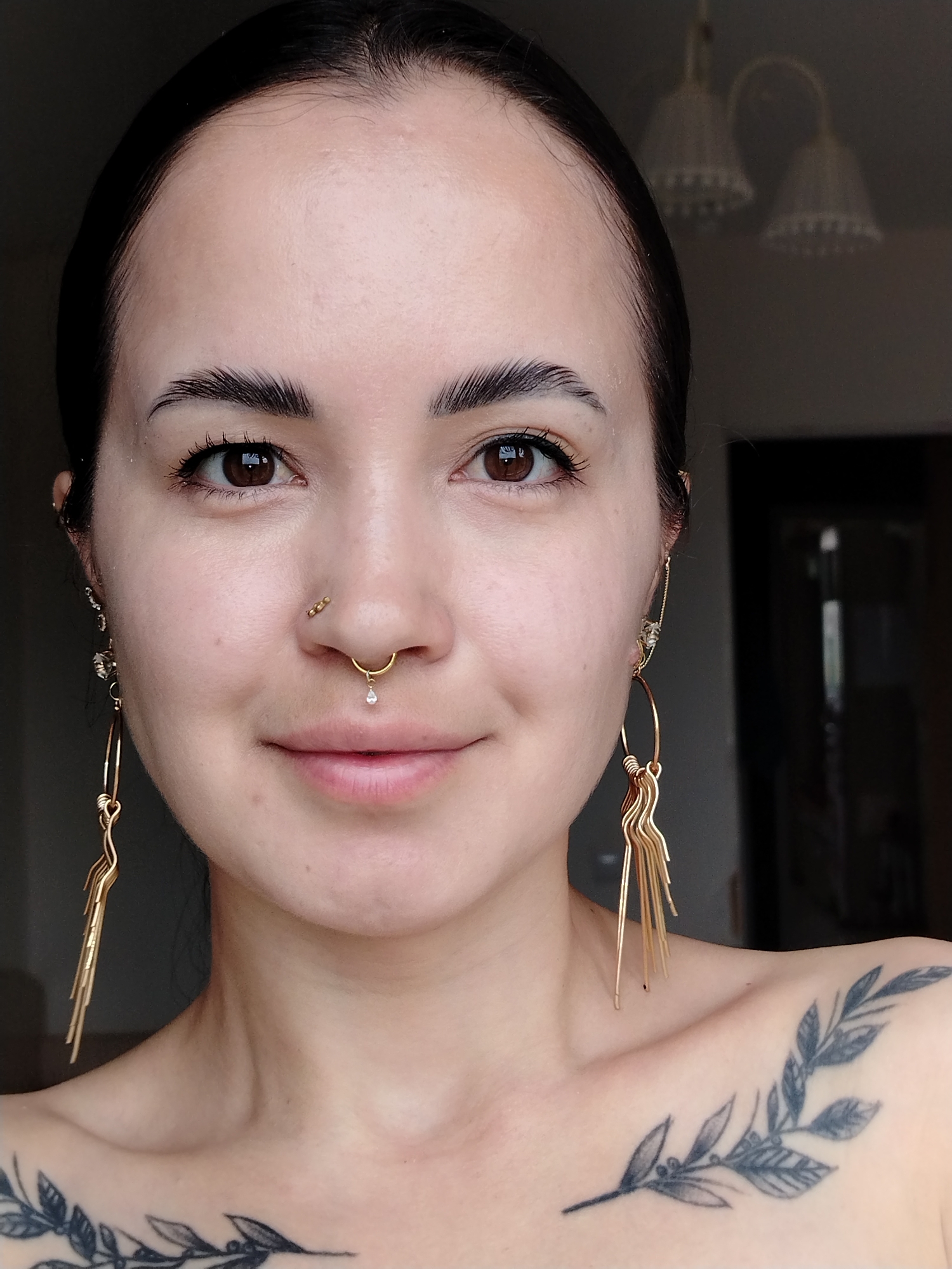 A medium shot captures a woman with fair skin, dark brown eyes, and dark hair pulled back. She has prominent eyebrows and several piercings: one in each nostril and a septum piercing. She wears dangling gold earrings. Her skin is clear with a few visible moles.
A tattoo of a leafy branch is visible on her chest and shoulder. The background is dimly lit and out of focus, showing a room interior with a hanging light fixture and a dark doorway. No specific information in the scene indicates its location in Noviy, Russia.
