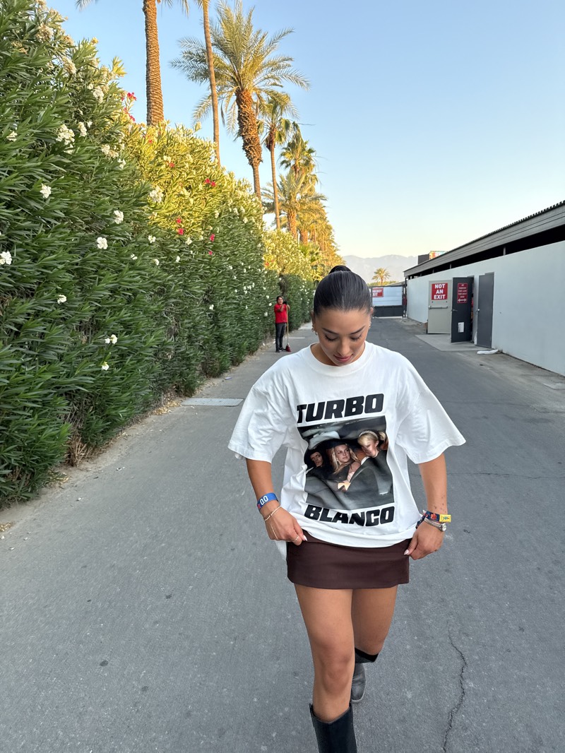 A young woman with her dark hair in a bun is standing on a street in Indio, California. She wears a white t-shirt with the words "TURBO BLANCO" and an image on the front, a short brown skirt, and black boots. She has bracelets on both wrists. She is looking down. Behind her is a tall green hedge with white flowers, palm trees, and a white wall. A man wearing a red shirt sweeps the street in the background. A sign that says "NOT AN EXIT" is visible on the white wall. The sky is light blue.