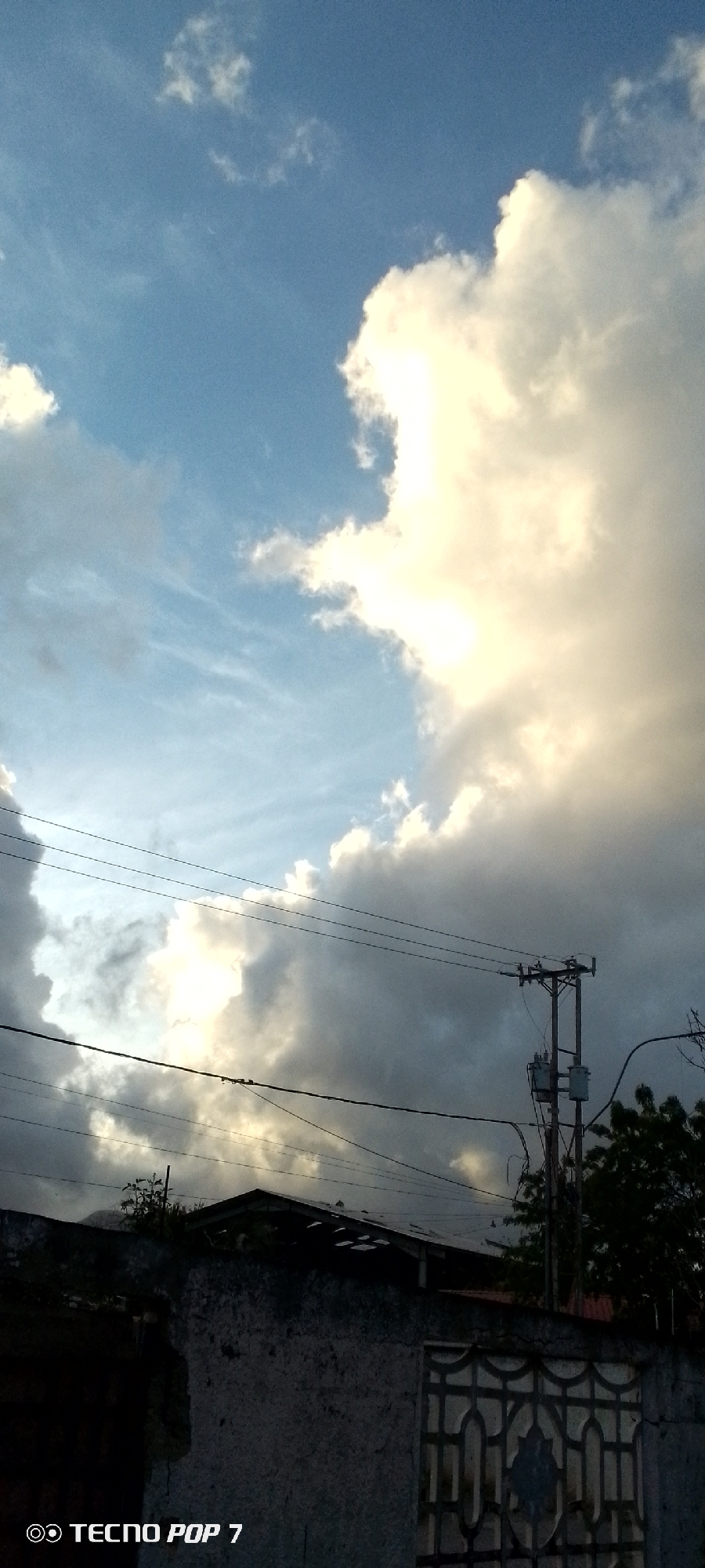 The image captures a view in Altagracia, Venezuela, showcasing a dynamic sky filled with clouds. The sky transitions from light blue to areas of dense, white cumulus clouds. In the foreground, a weathered concrete wall with a decorative metal grate provides a sense of enclosure. A building with a damaged roofline is visible behind the wall. Utility poles and wires traverse the scene, adding a layer of urban infrastructure. Trees and foliage partially obstruct the view of the building. The bottom left corner of the image features the inscription "TECNOP POP 7." The overall scene presents a contrast between the expansive sky and the elements of urban life in Altagracia.