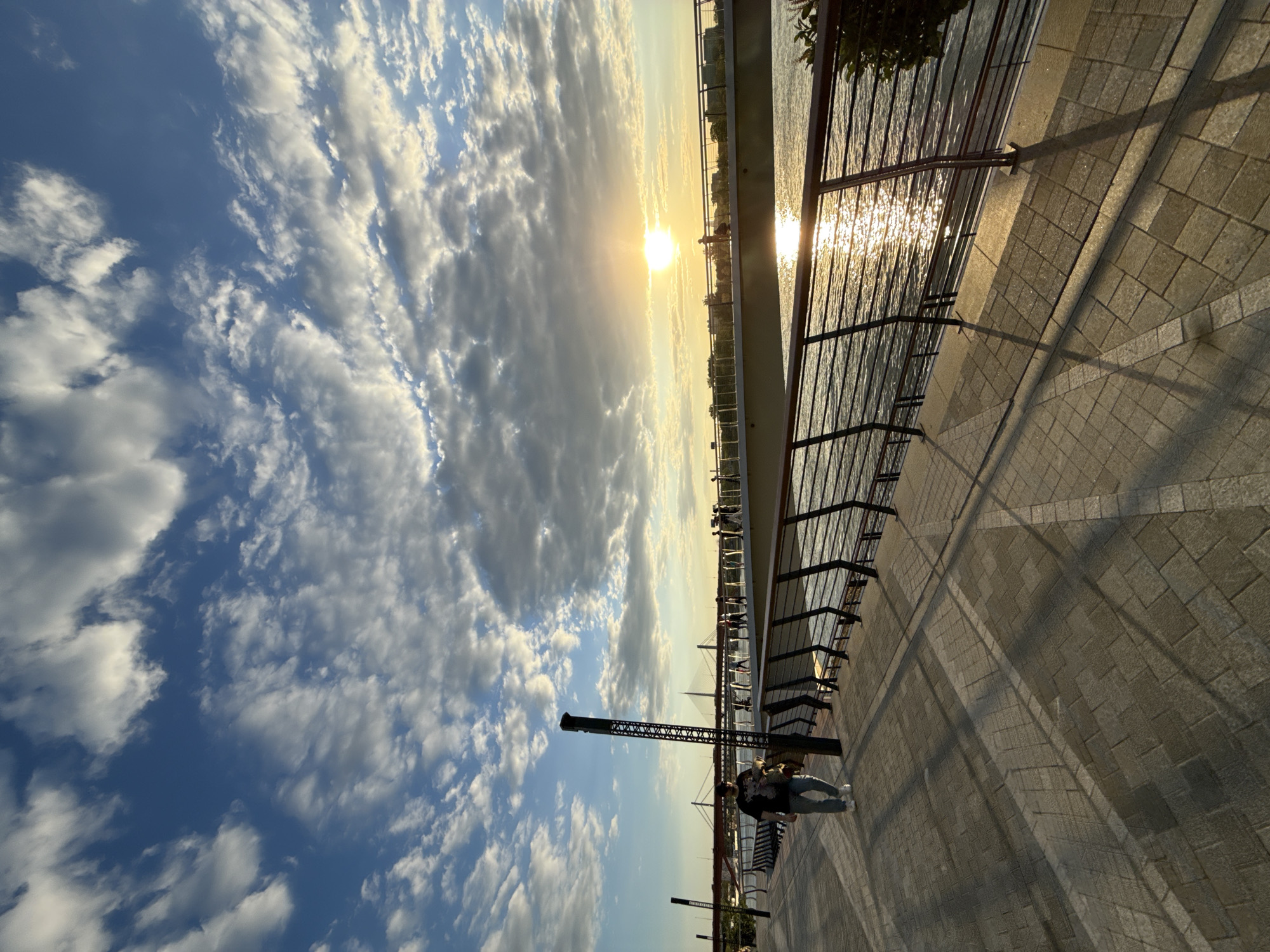 The image captures a picturesque sunset scene along a riverside promenade, likely in Belgrade, Serbia. The vibrant sun, a bright golden orb, is low on the horizon, dominating the upper right portion of the frame and casting an intense, shimmering reflection across the water to the right.
The sky is a prominent feature, showcasing a clear blue expanse adorned with a dynamic array of white and grey clouds that stretch horizontally across the frame. These clouds vary in density, with some appearing thin and wispy while others are more voluminous.
In the foreground, a paved walkway, constructed from light-colored rectangular blocks, extends into the distance. A dark metal railing separates the promenade from the expansive body of water. Long, sharp shadows, cast by the railing and other unseen objects, stretch diagonally across the walkway, emphasizing the low angle of the setting sun.
A single person is visible standing near the railing in the mid-ground, appearing to observe the scenic view. They are dressed in light blue jeans, white sneakers, and a dark patterned top. Further along the promenade, a tall, dark, lattice-like structure stands out. Across the water, the distant bank reveals the silhouettes of various buildings and what appears to be a bridge or significant architectural structure against the glowing sky. Dark foliage from trees or bushes is visible in the upper right corner, framing the scene. The overall ambiance is tranquil and serene, capturing the beauty of a sunset by the water. No visible text is present in the image.