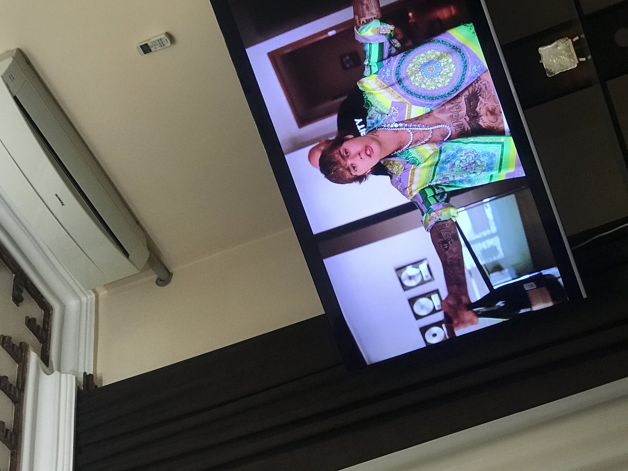 A television displays a man in Versace clothing, likely a music video. An air conditioning unit and a remote are attached to the wall nearby. The setting is indoors, possibly a residence. The location in Yerevan, Armenia cannot be determined.