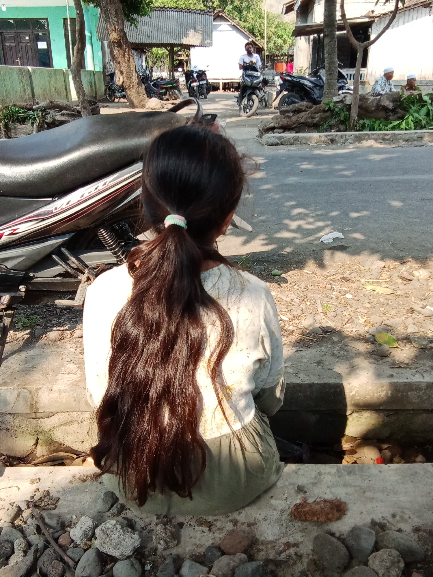 A young girl with long, dark hair tied in a ponytail sits on a concrete ledge, facing away from the camera. She is wearing a light-colored shirt with a floral pattern and a green skirt. To her left, a portion of a black motorcycle is visible. The background depicts a street scene with several other motorcycles parked. In the distance, buildings and trees are present. Two men, one wearing a white shirt and cap and the other in a patterned shirt and cap, are seated on the right side of the frame, with some foliage in front of them. The setting appears to be a street in Trebungan, Indonesia, characterized by its tropical climate with dappled sunlight creating shadows on the ground. The ground is covered with stones and debris, suggesting a rural or less developed urban area.
