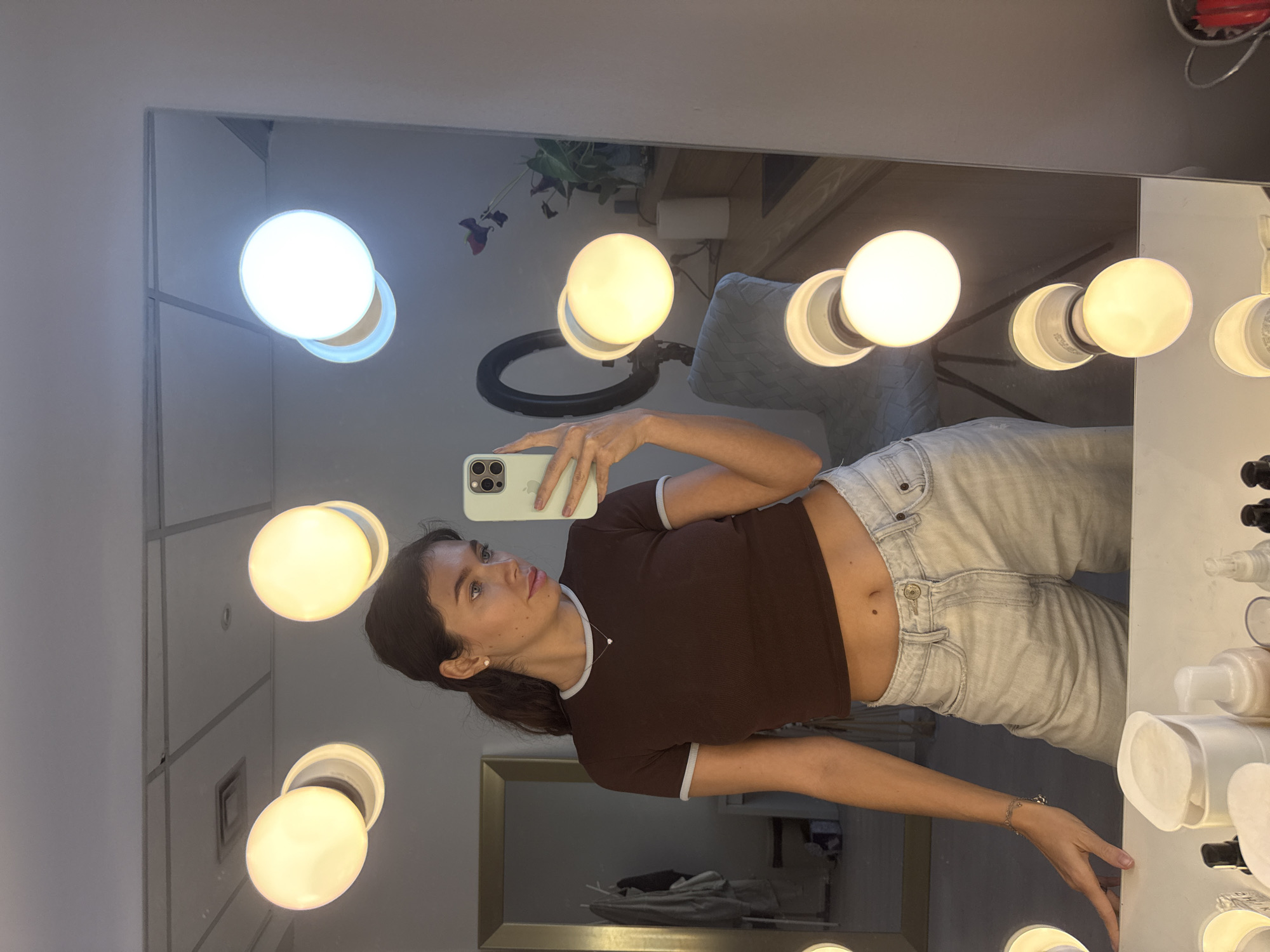 A young woman with long dark hair is taking a selfie in a vanity mirror in what appears to be Ramat Gan, Israel. The mirror is lined with multiple light bulbs, and there are beauty products visible on the right side. The woman is wearing a brown t-shirt and light-wash jeans, and holding a light green iPhone. Reflected in the mirror are other room details, including a plant, a ring light, and furniture. Through another doorway in the back is another room.