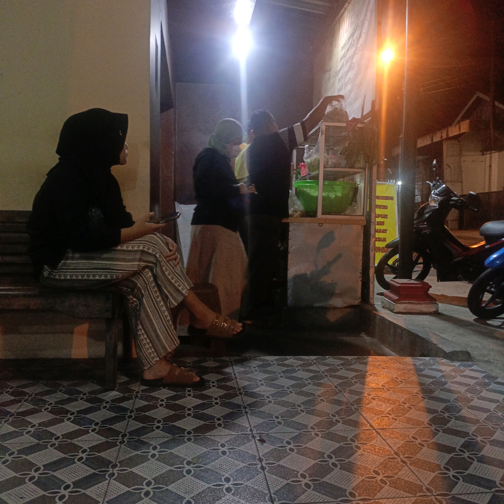 This image depicts a nighttime scene at a street-side food stall in Salatiga, Indonesia. In the foreground, a woman wearing a black hijab and patterned pants sits on a wooden bench, holding a smartphone. The floor is tiled with a geometric pattern. In the background, two individuals appear to be working at the stall, which is illuminated by bright overhead lights and a street lamp casting an orange glow. A green plastic basket and various items are visible on a shelf behind the stall. Two motorcycles, one black and one blue, are parked to the right of the stall. The overall atmosphere suggests a bustling, late-night local eatery.