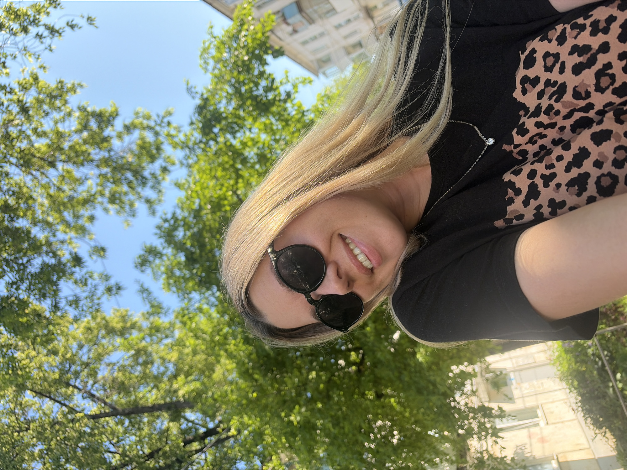 A young woman with blonde hair and sunglasses is taking a selfie outdoors. She is wearing a black t-shirt with a leopard print design and a delicate silver necklace. The background is filled with lush green trees and a clear blue sky, suggesting a sunny day. In the distance, buildings can be seen, indicating an urban or suburban setting. This scene is likely located in the city of Chaikovsky, Russia, a place known for its natural beauty and proximity to the Kama River.