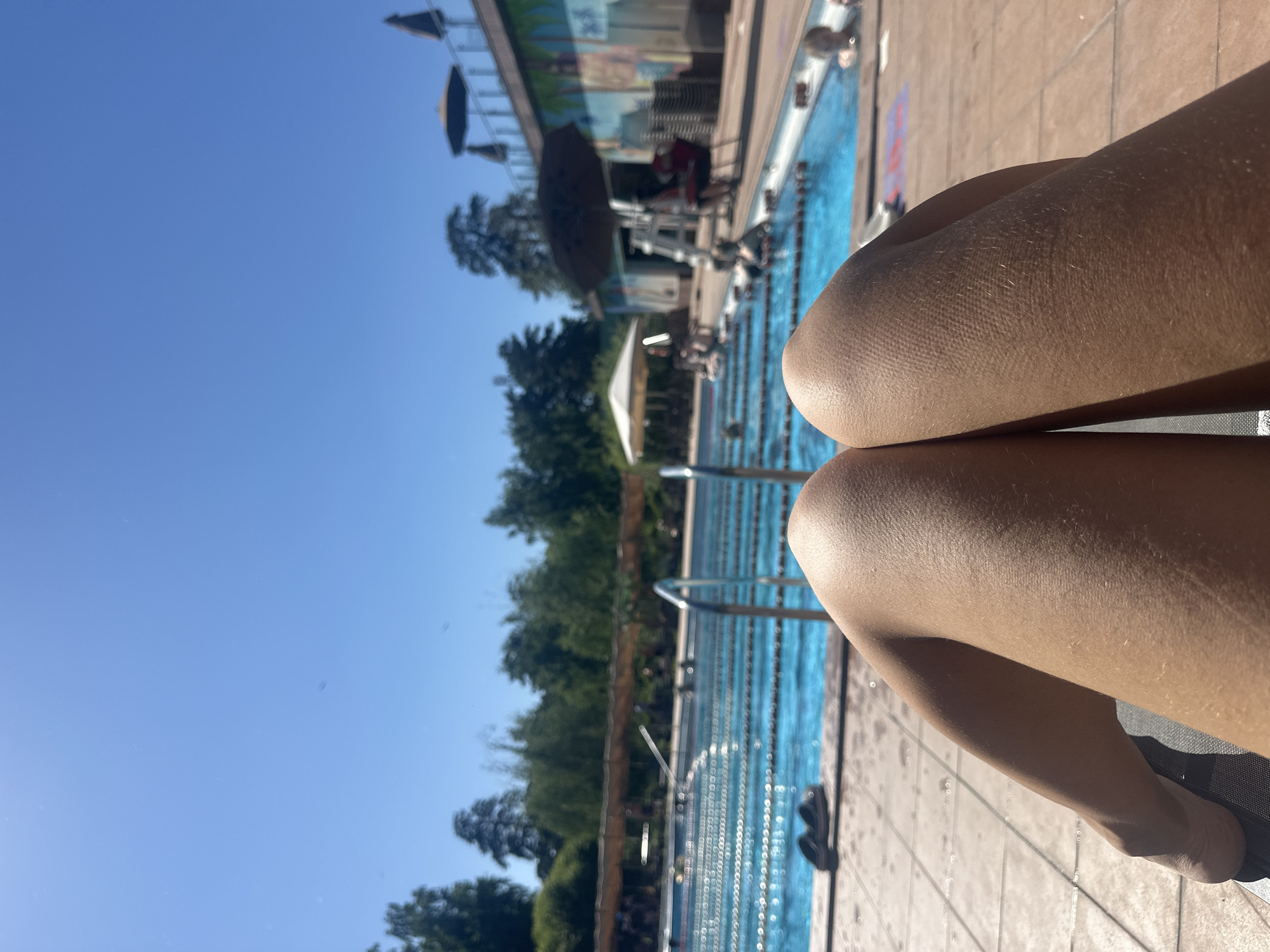 The image depicts a sunny day at an outdoor swimming pool. In the foreground, a person's legs are visible, resting on a lounge chair. The legs appear tan and are covered in fine hairs. The individual is wearing dark footwear. Beyond the foreground, a blue swimming pool is filled with water and divided into lanes by ropes. Several people are swimming or wading in the pool. In the background, there are trees, a building with a mural, umbrellas, and lounge chairs. The sky is clear and blue. The location is identified as the city of Berdsk municipality, Russia.