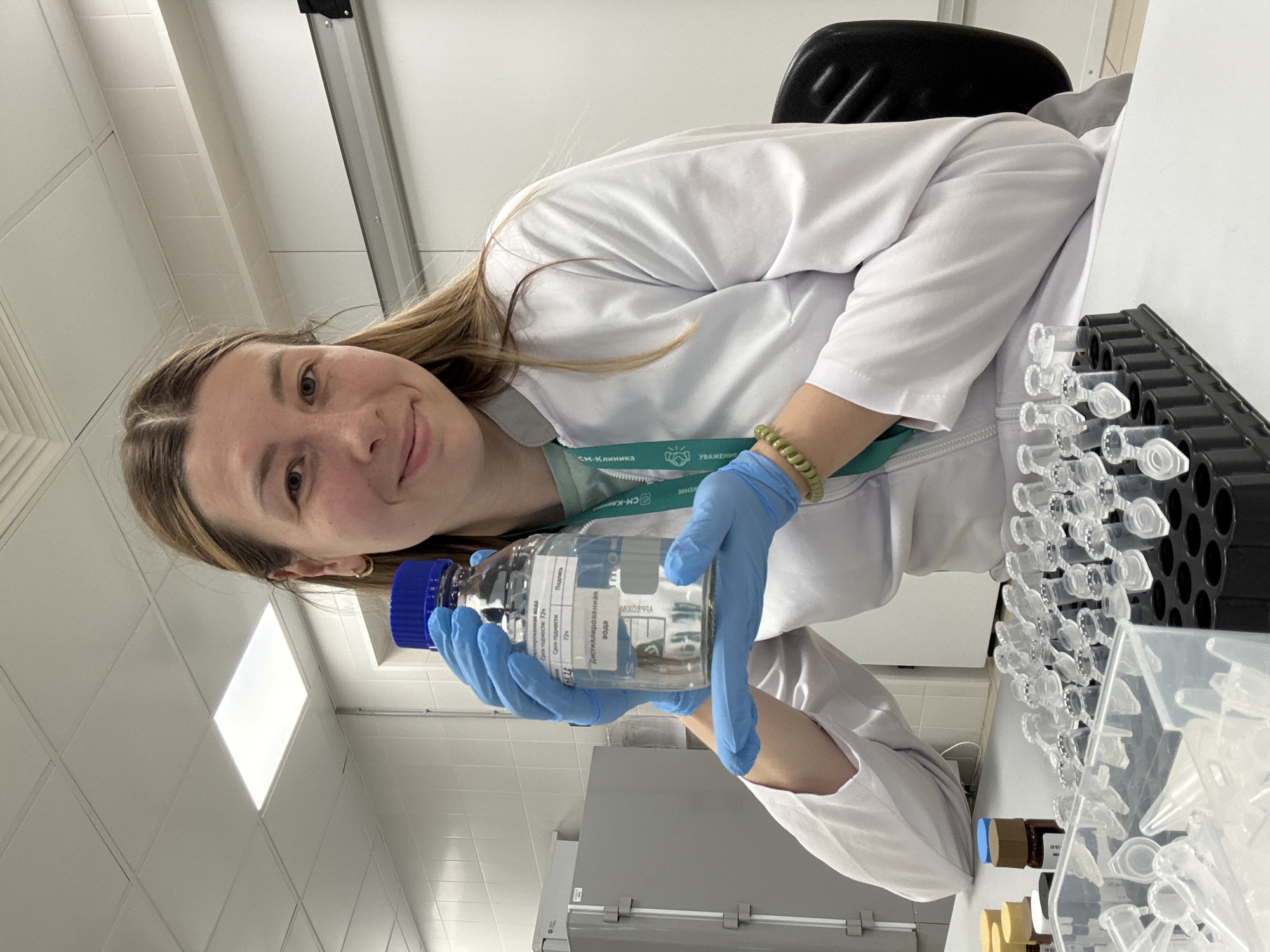A woman in a white lab coat and blue gloves is holding a bottle of distilled water. The bottle's label is partially in Russian. In the background, there is a rack of test tubes. The woman is wearing a green lanyard with the words "СМ-Клиника" and "Уважение" written on it, indicating a connection to the "CM-Clinica" (SM-Clinic) network. This may be located in Moscow, Russia, based on the Russian text on the label and lanyard.