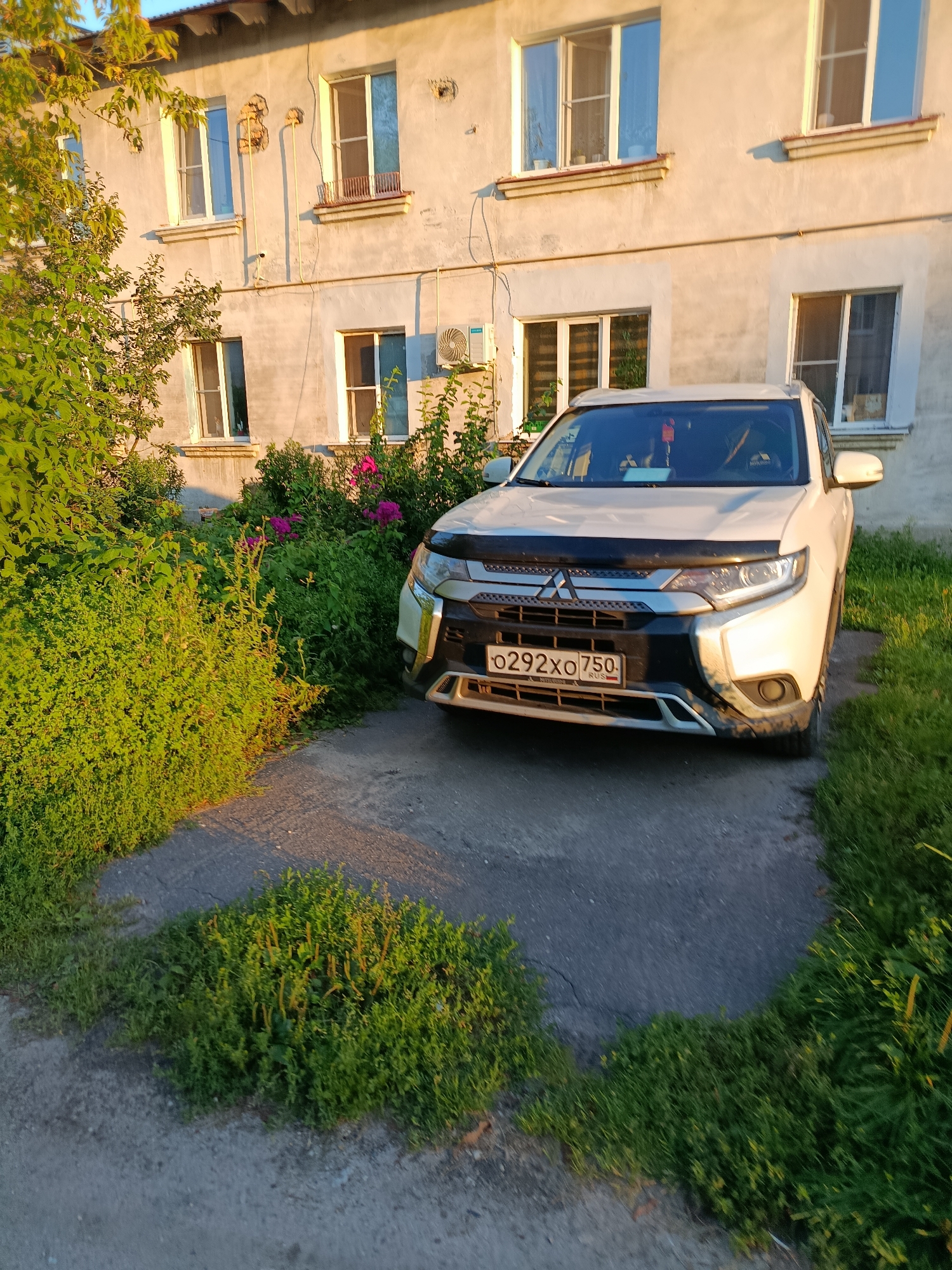 The image depicts a white Mitsubishi Outlander parked on a concrete patch in front of a weathered, multi-story building. The building has several windows with white frames, some of which appear to have grates or decorative elements. The exterior wall of the building is painted a light color, showing signs of wear and tear. Lush green foliage, including bushes and trees, surrounds the parking area and climbs up the side of the building. There are also visible pink flowers among the greenery. The license plate of the car is clearly visible and reads "0292XO 750 RUS". The scene appears to be set in a residential area, likely in the city of Vereya, Russia, given the context. The lighting suggests it is either early morning or late afternoon, with the sun casting shadows and highlighting the textures of the building and vegetation. There are no people visible in the image.