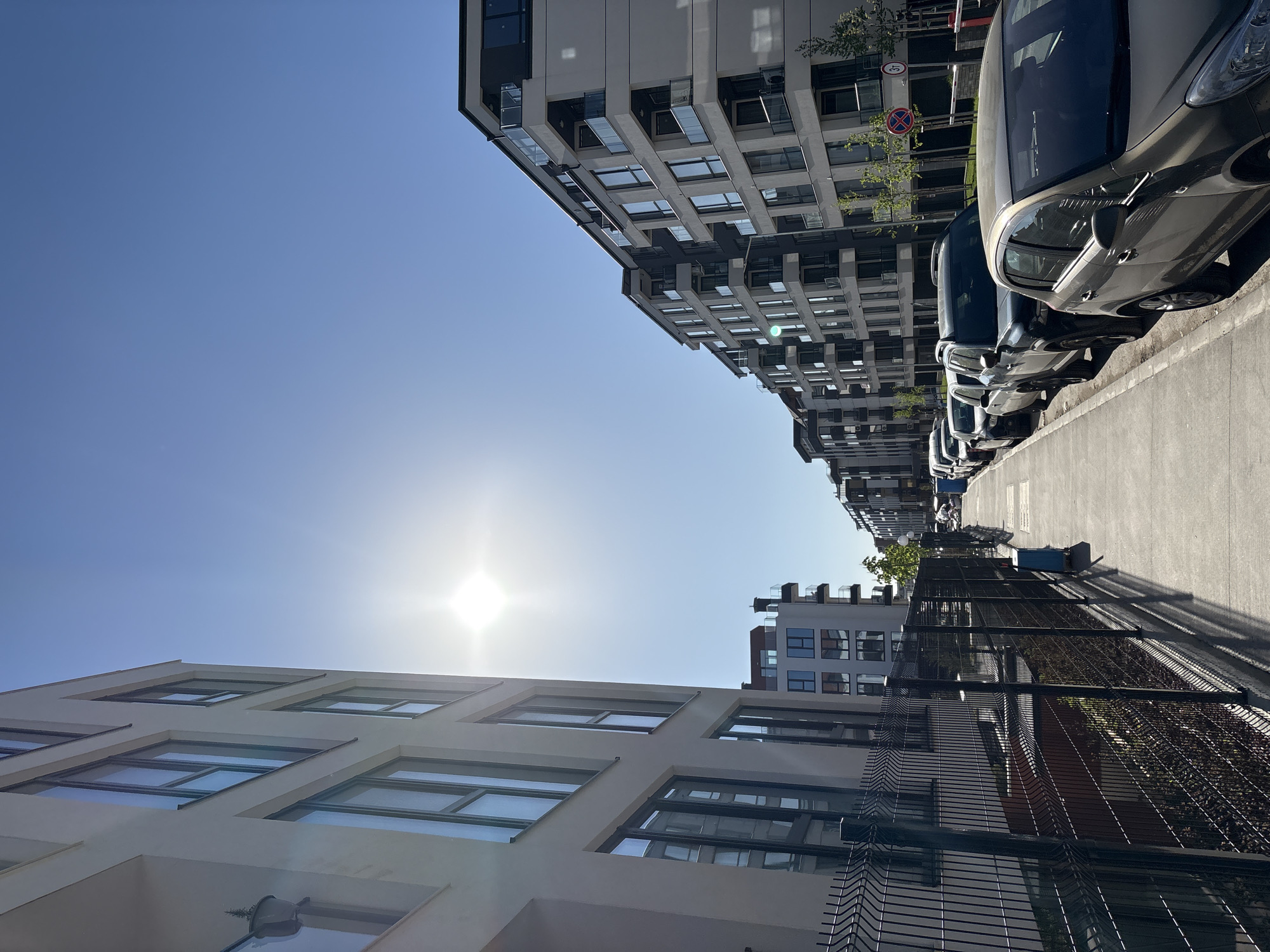 The image shows an urban street scene in Sofia, Bulgaria, taken on a sunny day. Modern buildings with repetitive window patterns line both sides of the street. Parked cars are visible on the right side of the road. A metal fence runs along the bottom right of the frame. The sun is high in the sky, creating a glare. The image appears to be taken looking upwards along the street.